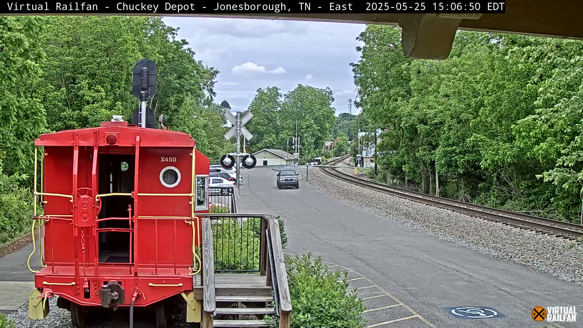 A red railroad car is parked in front of a rail crossing, with a partly cloudy sky and lush green trees surrounding a paved road and railroad tracks.