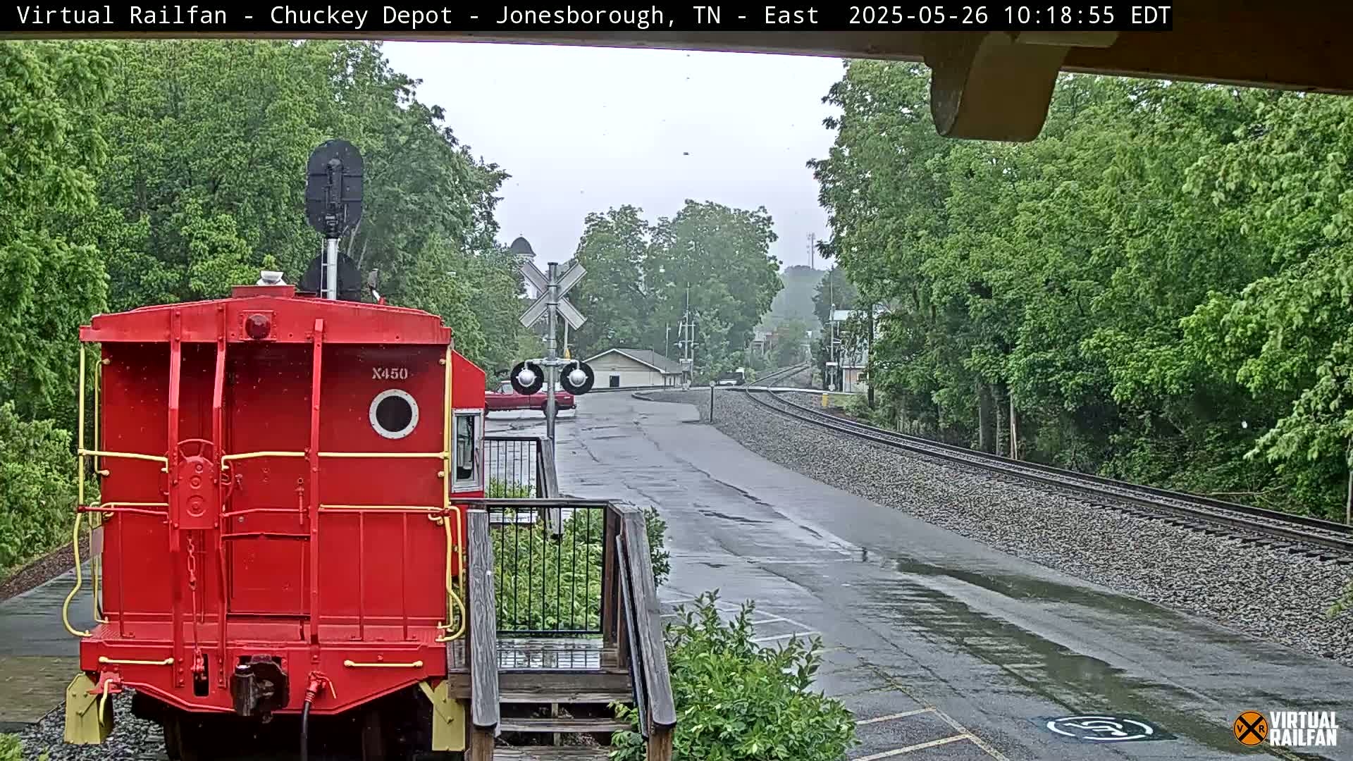 A red railway car is parked near a railway crossing, with a wet road and lush green trees visible in the background under an overcast sky.