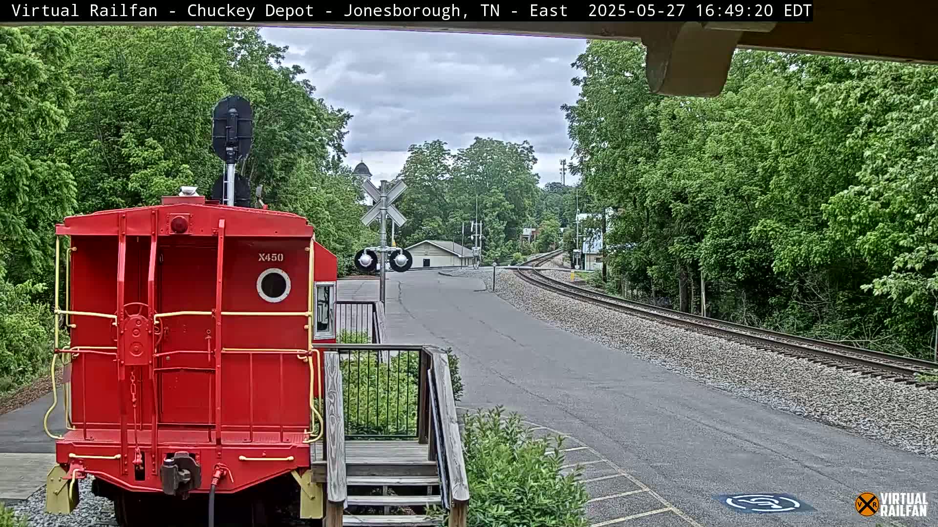 A red railroad car is parked at a train depot on a partly cloudy day, with railroad tracks extending into the distance, surrounded by lush green trees.
