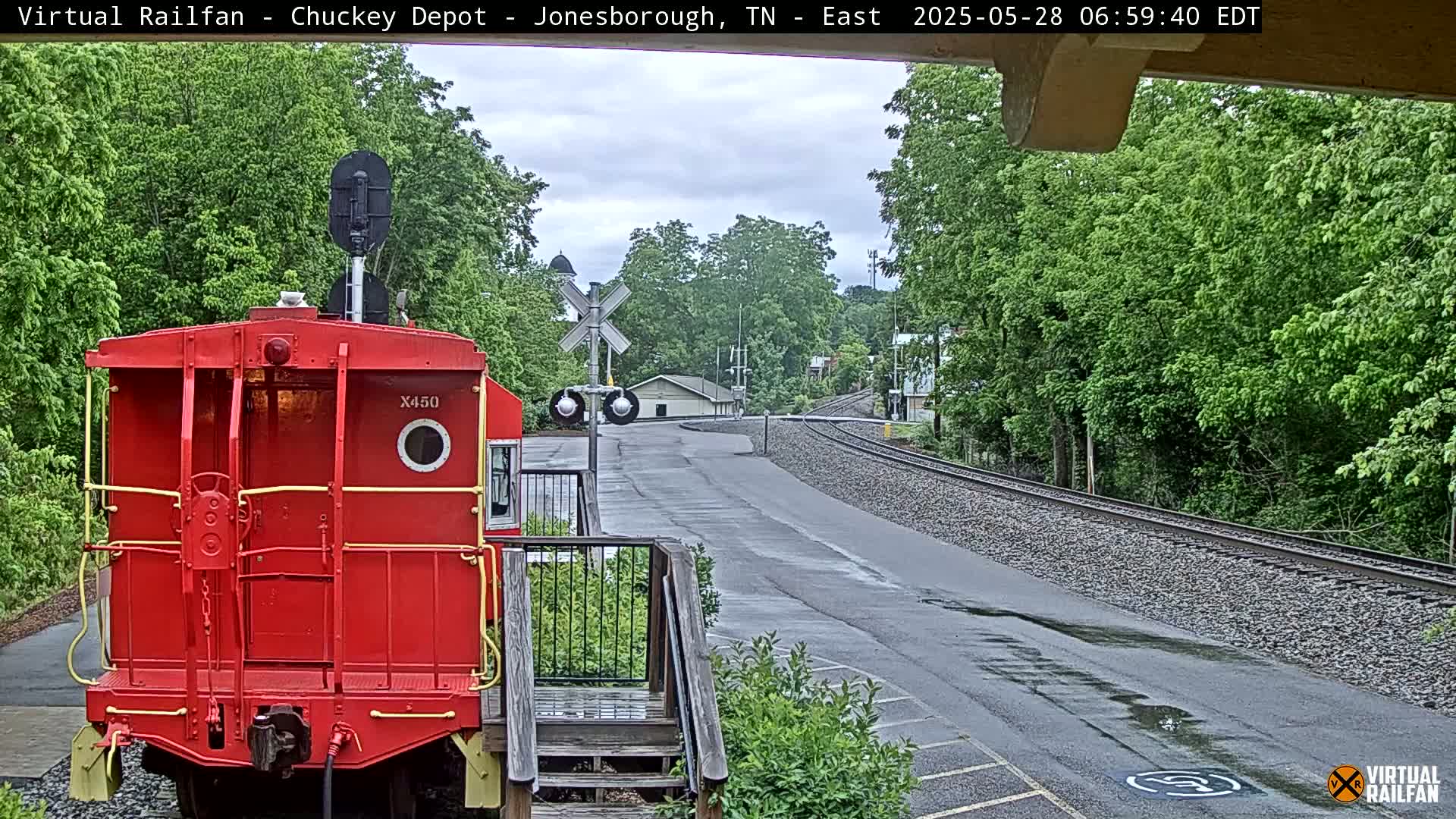 A red railcar is parked near a railway crossing on a wet road, with lush green trees and overcast skies.