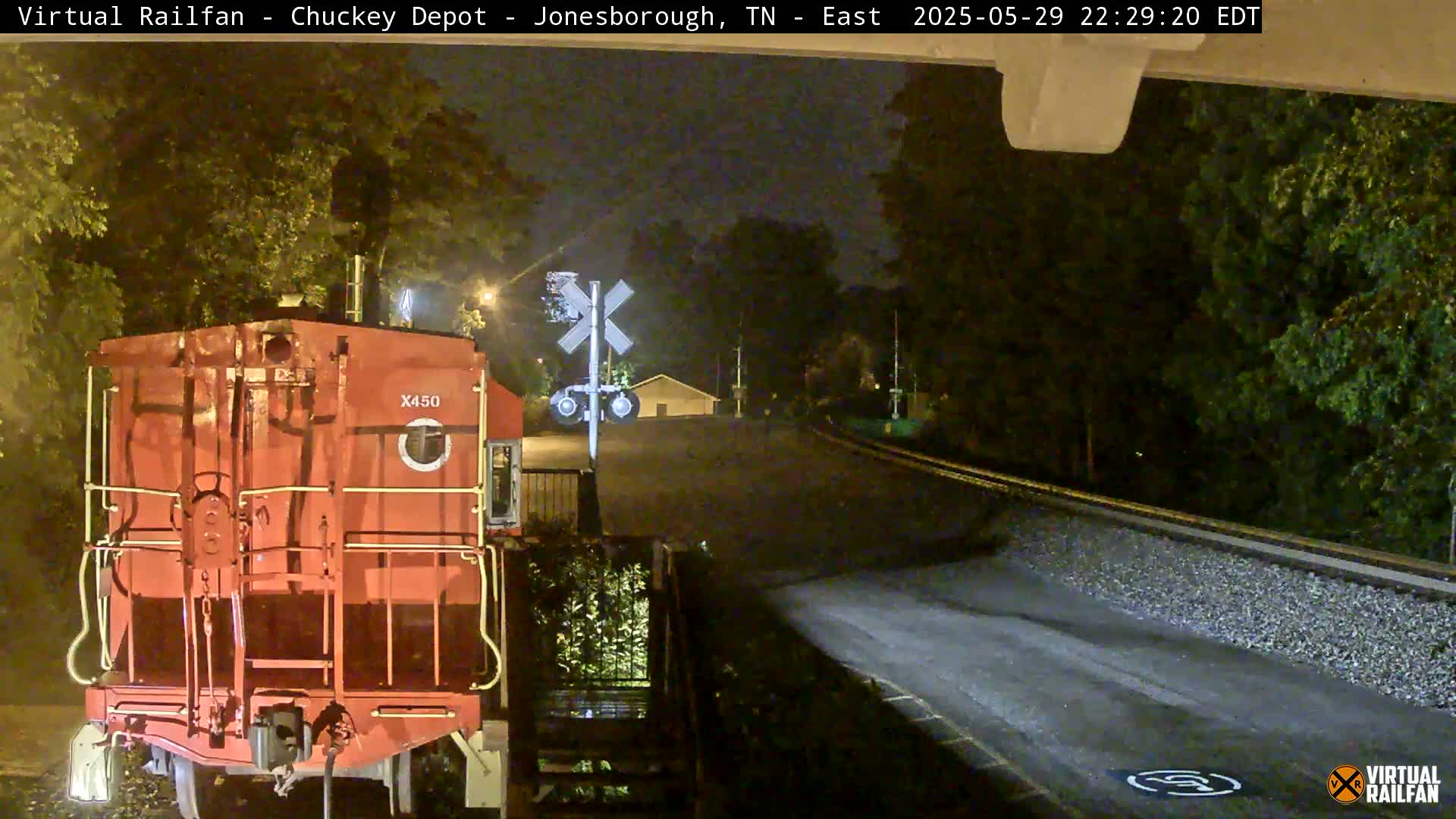 At night, a red railroad car sits at a railroad crossing near trees under a dark sky.
