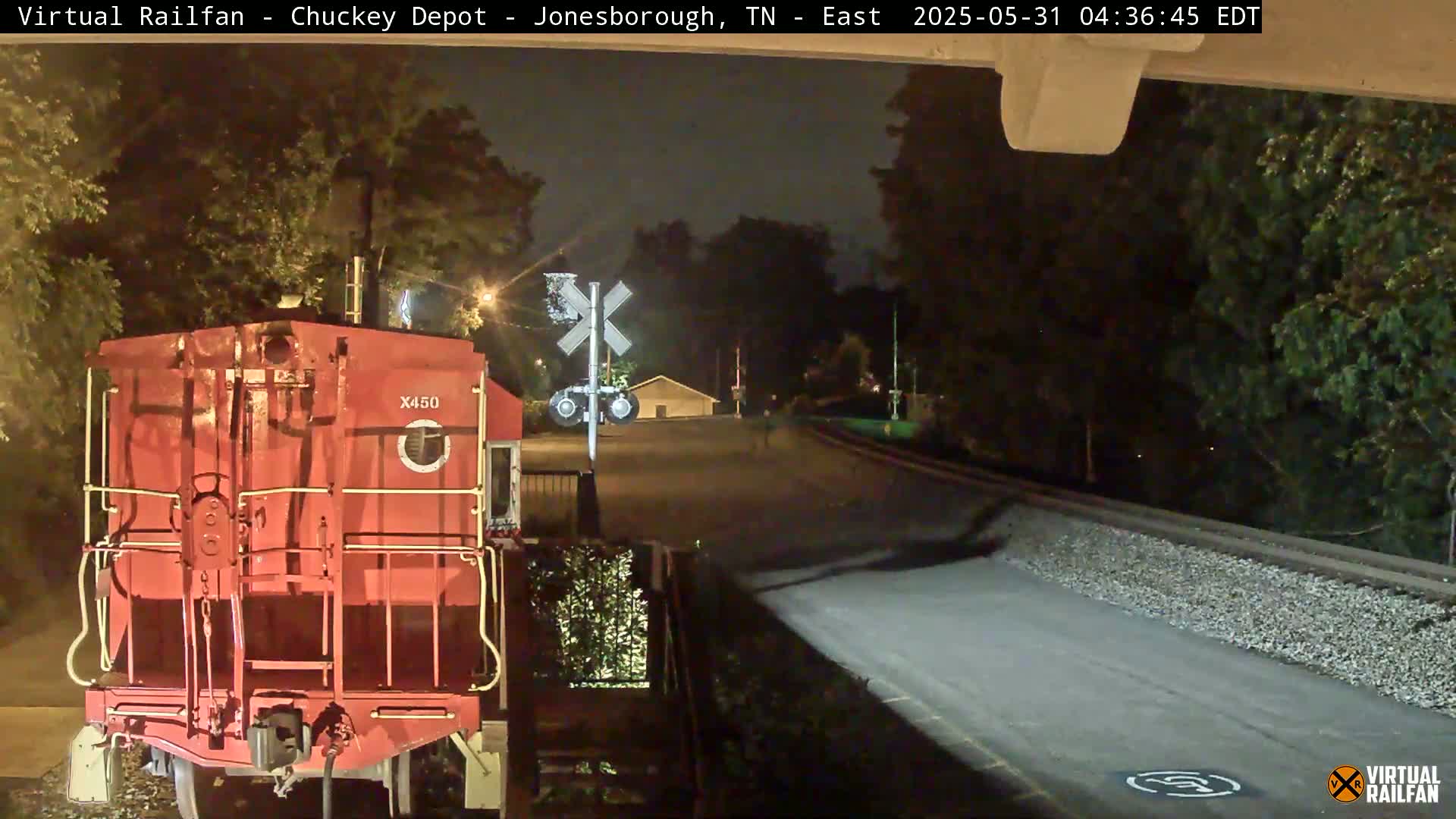At night, a red train car is positioned in front of a railroad crossing near a road, with trees lining the background.