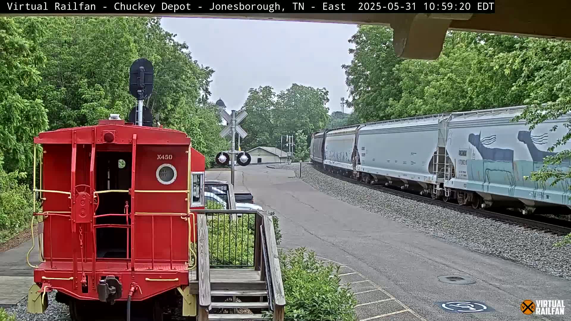 A long freight train passes a red caboose on a partly cloudy day, with lush green trees in the background.