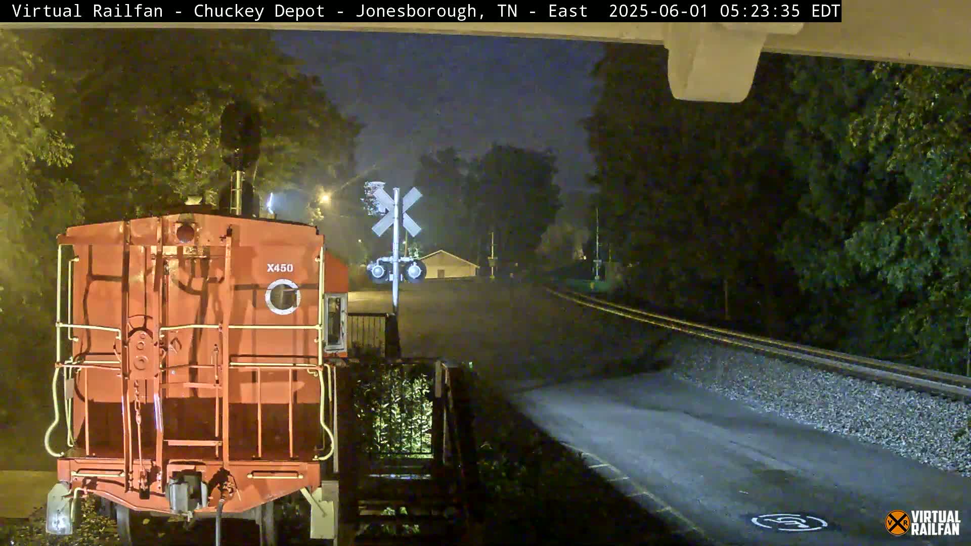 At night, a red train car sits at a railroad crossing near some trees.