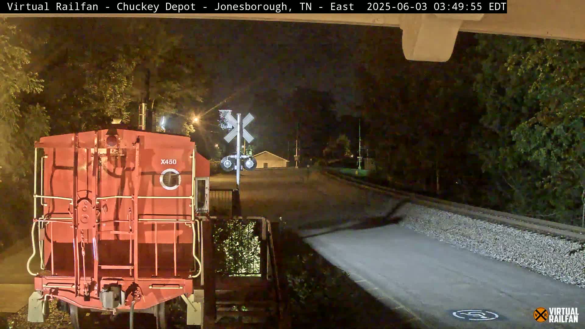 A red train car is stopped at a nighttime railroad crossing near some trees under a dark sky.