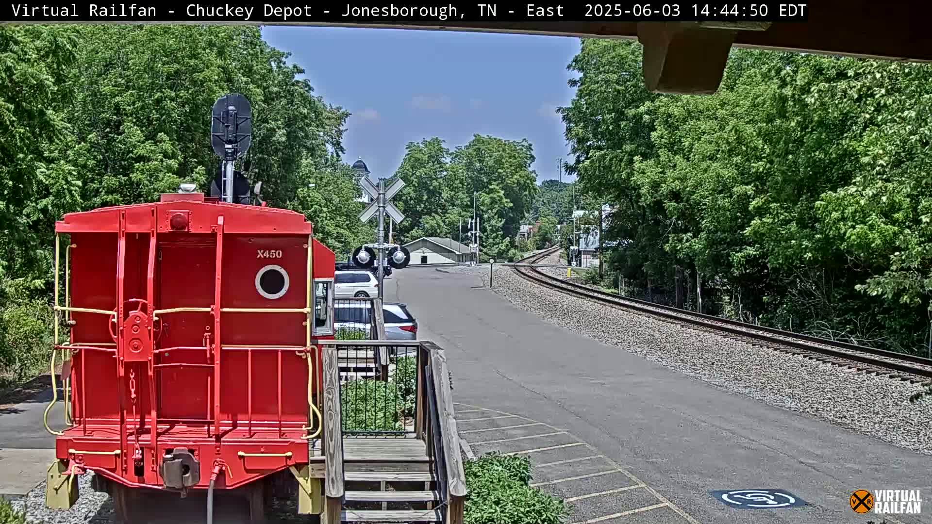 A red train car is parked facing the camera, with a rail yard and road leading towards a town under a clear sky.