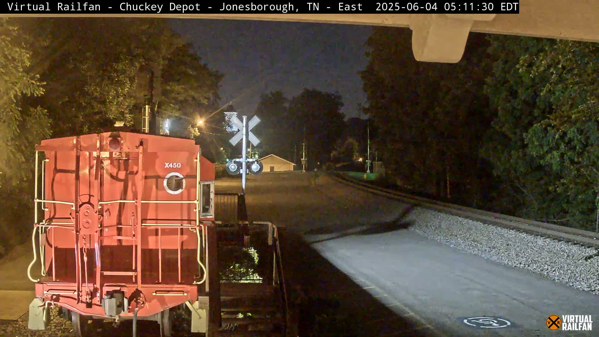 At night, a red railroad car sits at a train crossing near a road with trees and a small building in the background.