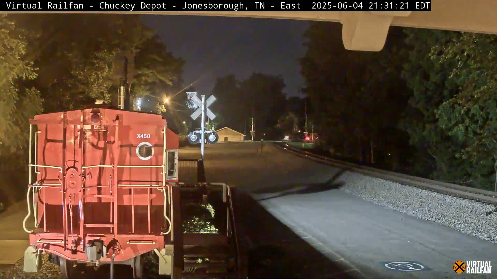 A red railroad car is stopped at a nighttime crossing, with trees and a road visible in the background.