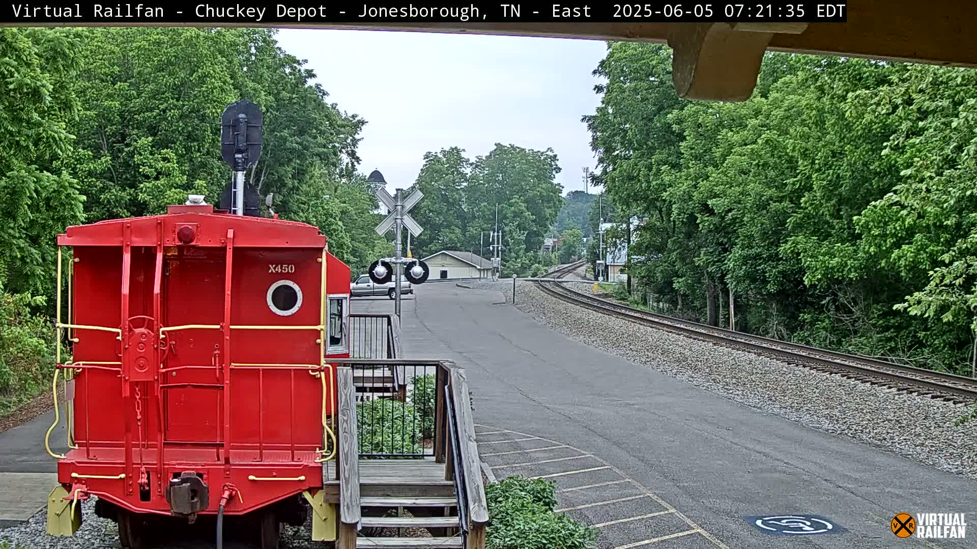 A red railroad car is parked at a train depot on a partly cloudy day, with railroad tracks curving away into a wooded area.