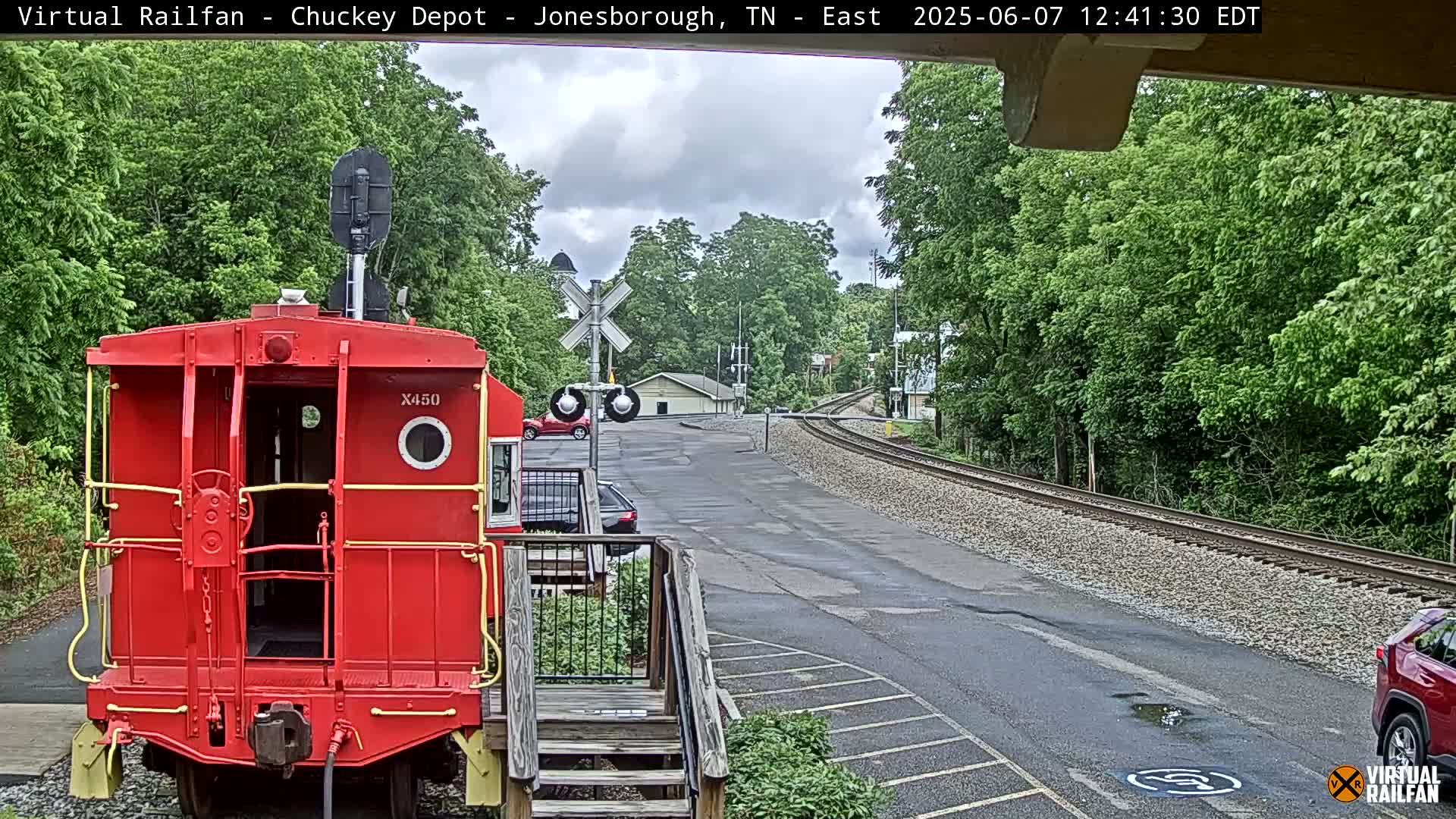 A red caboose sits in the foreground of a railroad crossing under a partly cloudy sky, with a curving railway track and some cars parked nearby.