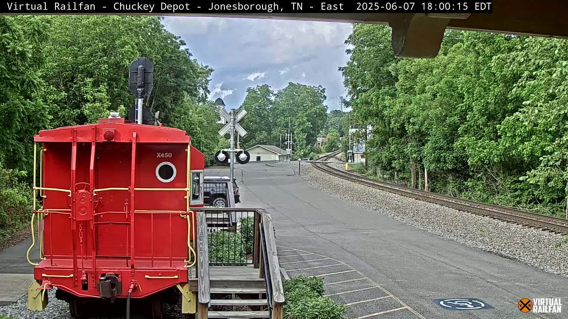 A red railroad car is parked at a rail yard on an overcast day, with a paved road leading to a railroad crossing and lush green trees surrounding the area.