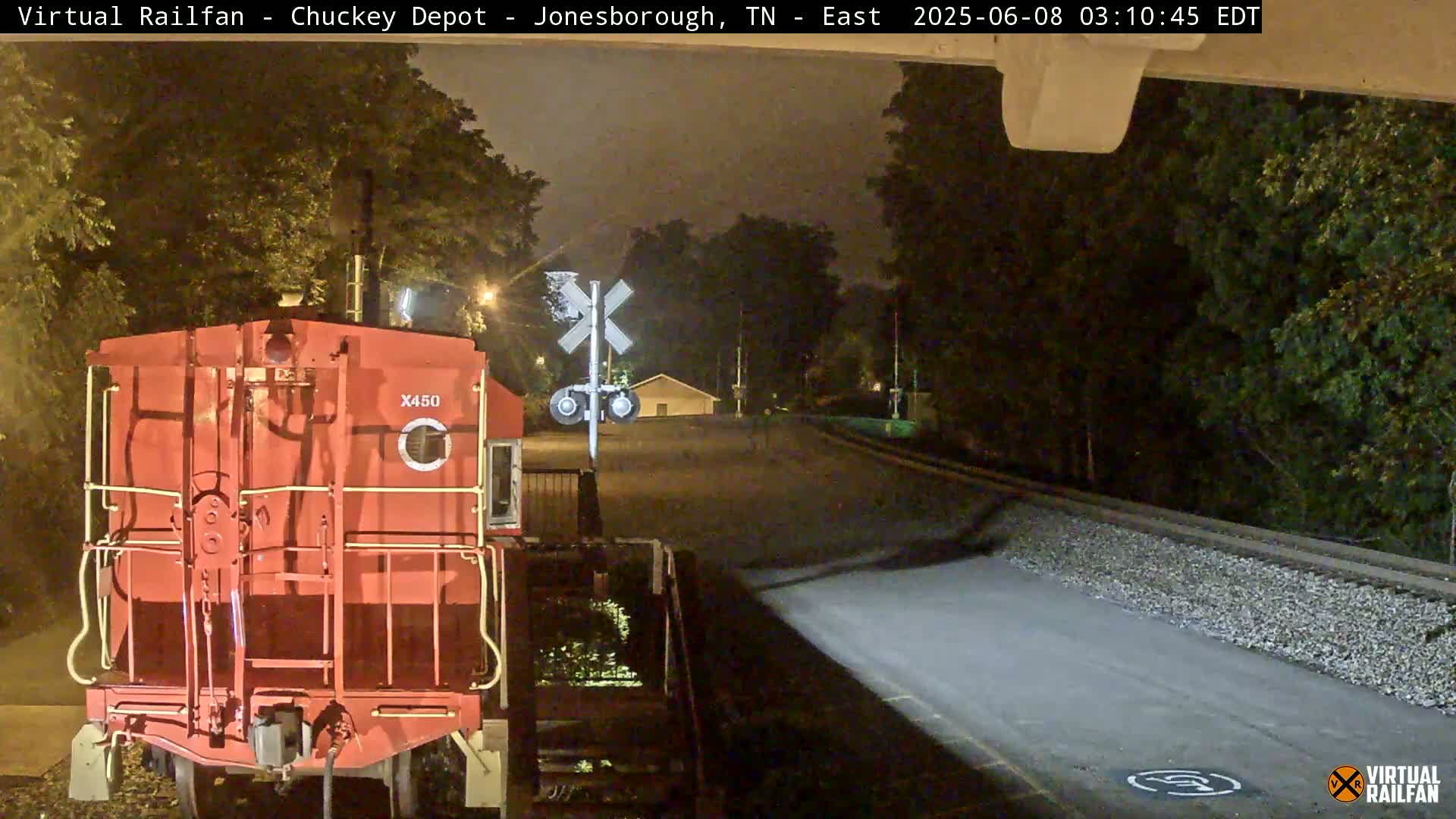 A red railroad car is parked at night near a railroad crossing under a dark, overcast sky.