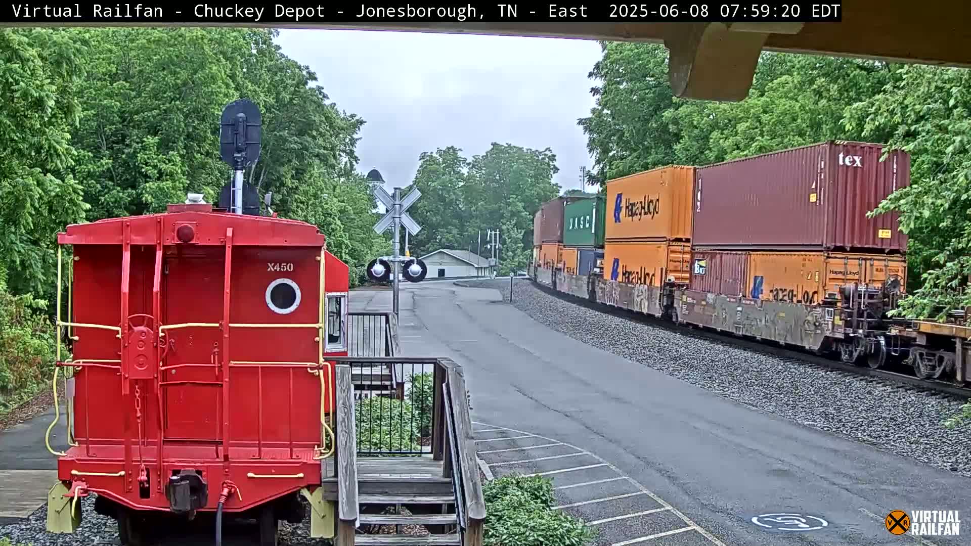 A red railcar is in the foreground, facing a train carrying multiple shipping containers that is passing by under an overcast sky.