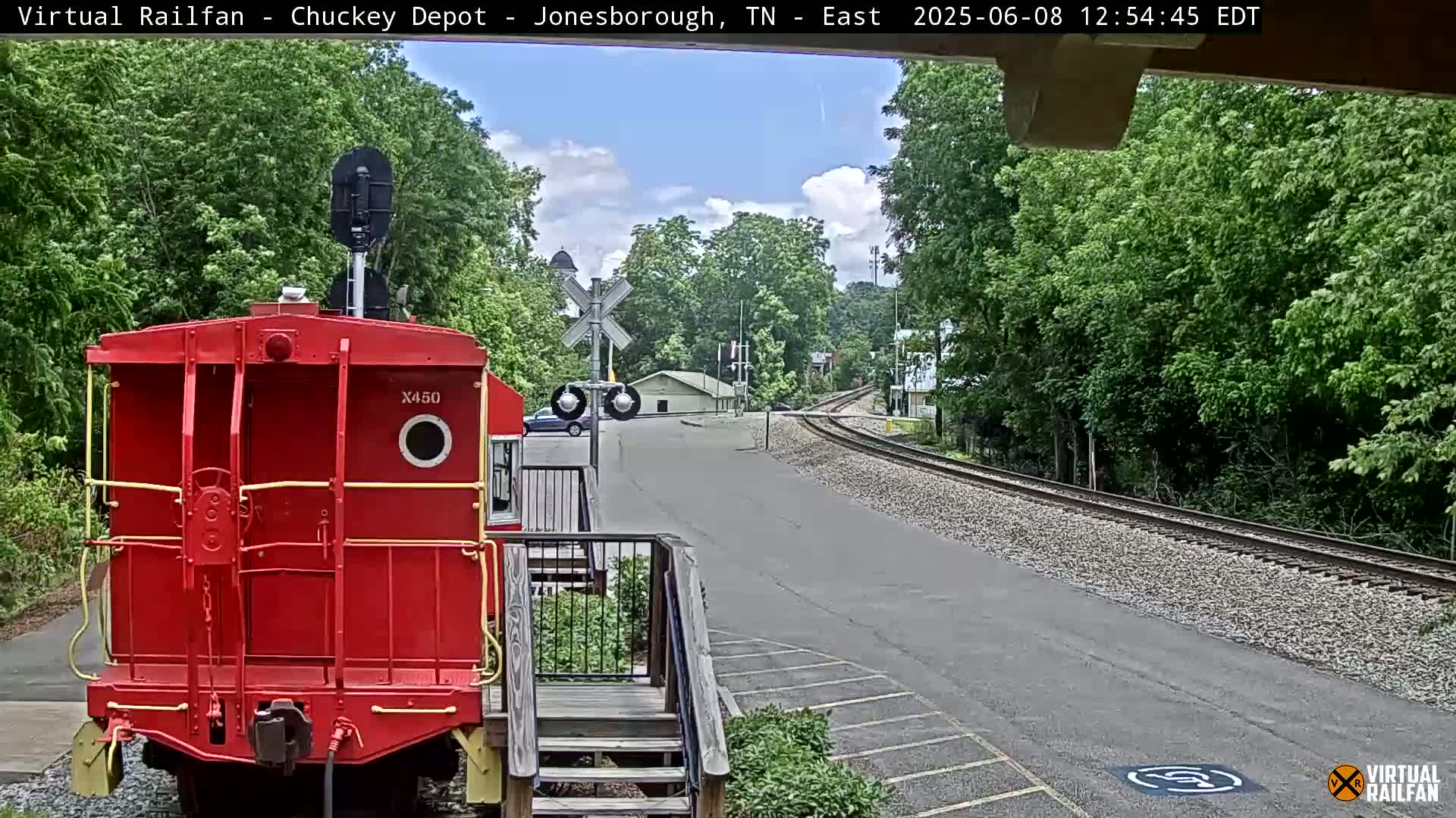 A red railroad car is parked at a train depot on a partly sunny day, with railroad tracks curving into the distance, surrounded by lush green trees.