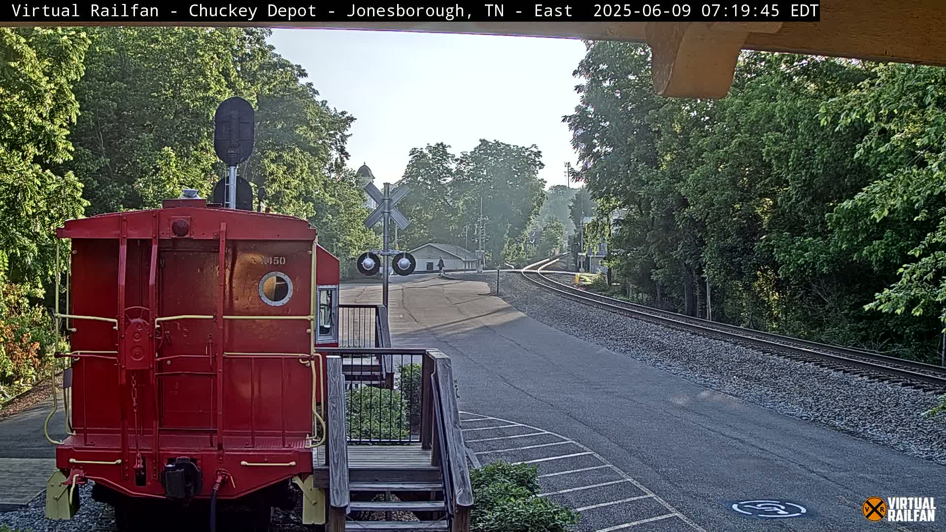 A red caboose sits on a railway track next to a paved road, with lush green trees and a partly cloudy sky in the background.