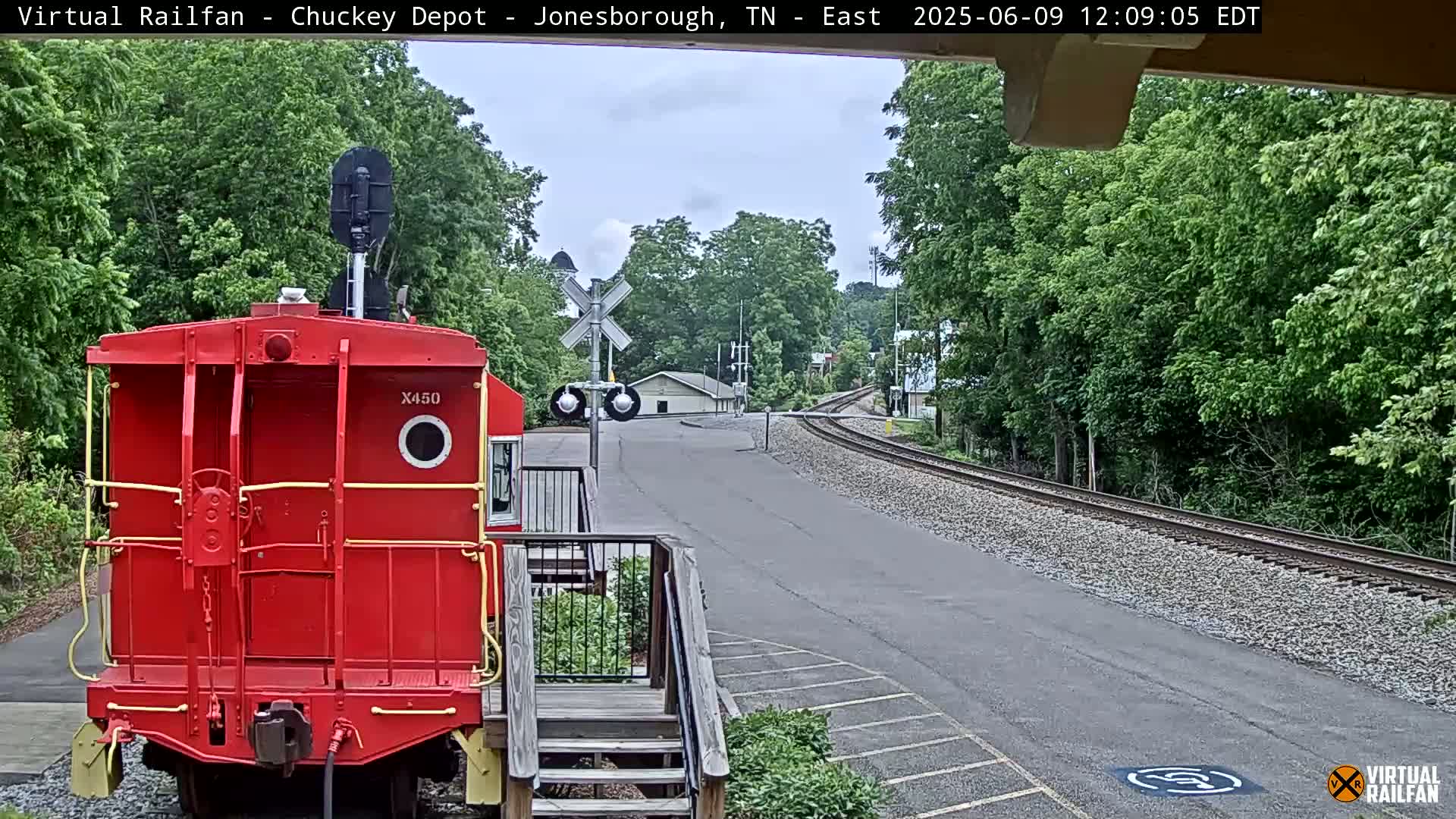 A red railway car is parked near a railroad crossing on a mostly sunny day, with green trees in the background.