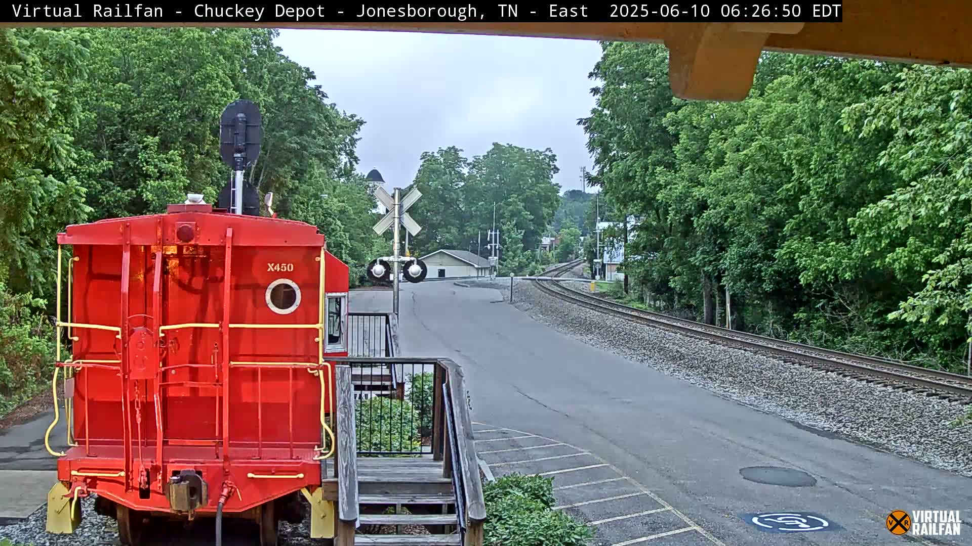 A red train car is parked facing away from the camera, next to a railroad track that curves away into a tree-lined area under an overcast sky.