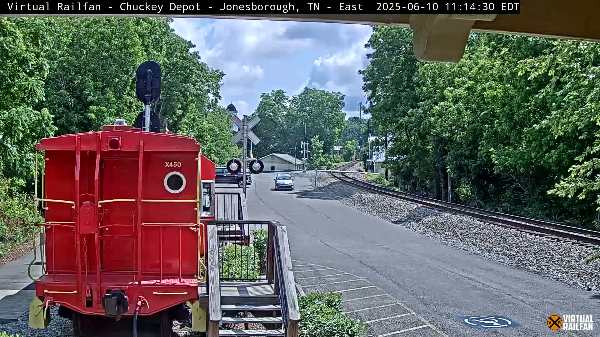A red train car is parked at a railroad crossing on a sunny day, with a car driving past on a nearby road.