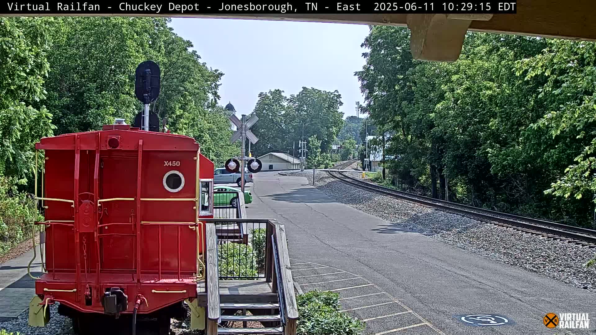 A red railroad car is parked at a train depot on a sunny day, with railroad tracks extending into the distance beside a paved road lined with green trees.