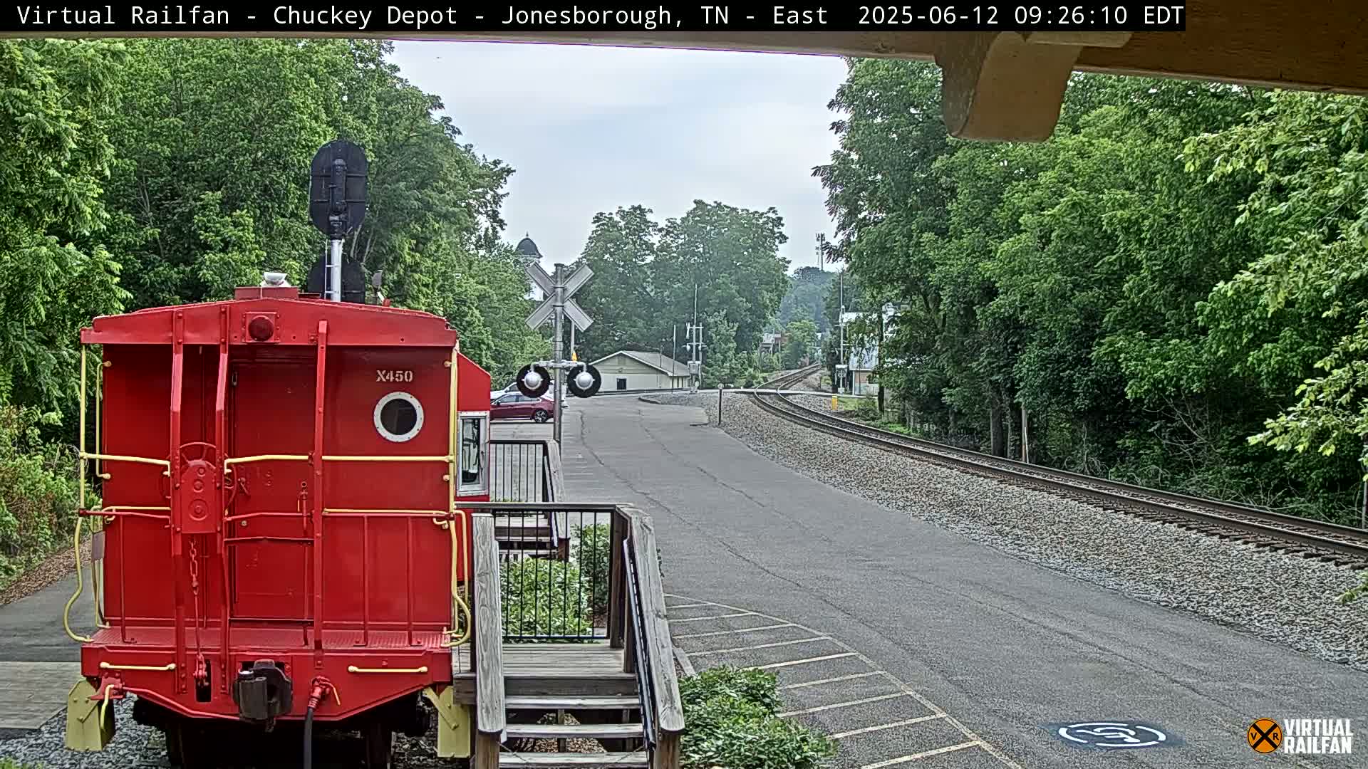 A red railroad car is parked next to a paved road and railway tracks, under an overcast sky.