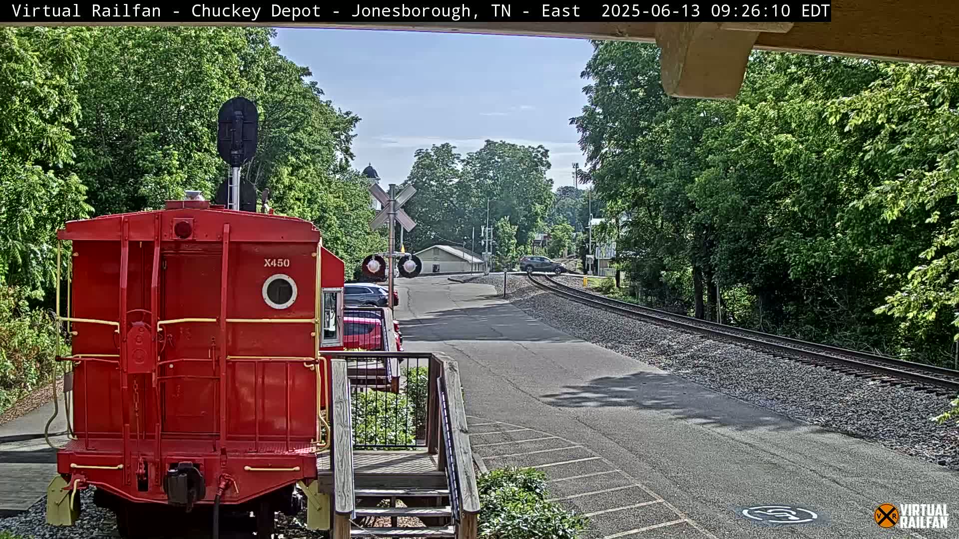 A red railway caboose is parked near a roadway next to train tracks on a sunny day, with lush green trees lining the background.