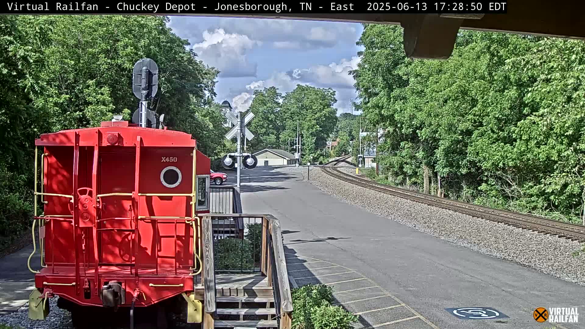 A red train car sits in front of a train track that leads to a small building, all under a partly cloudy sky.