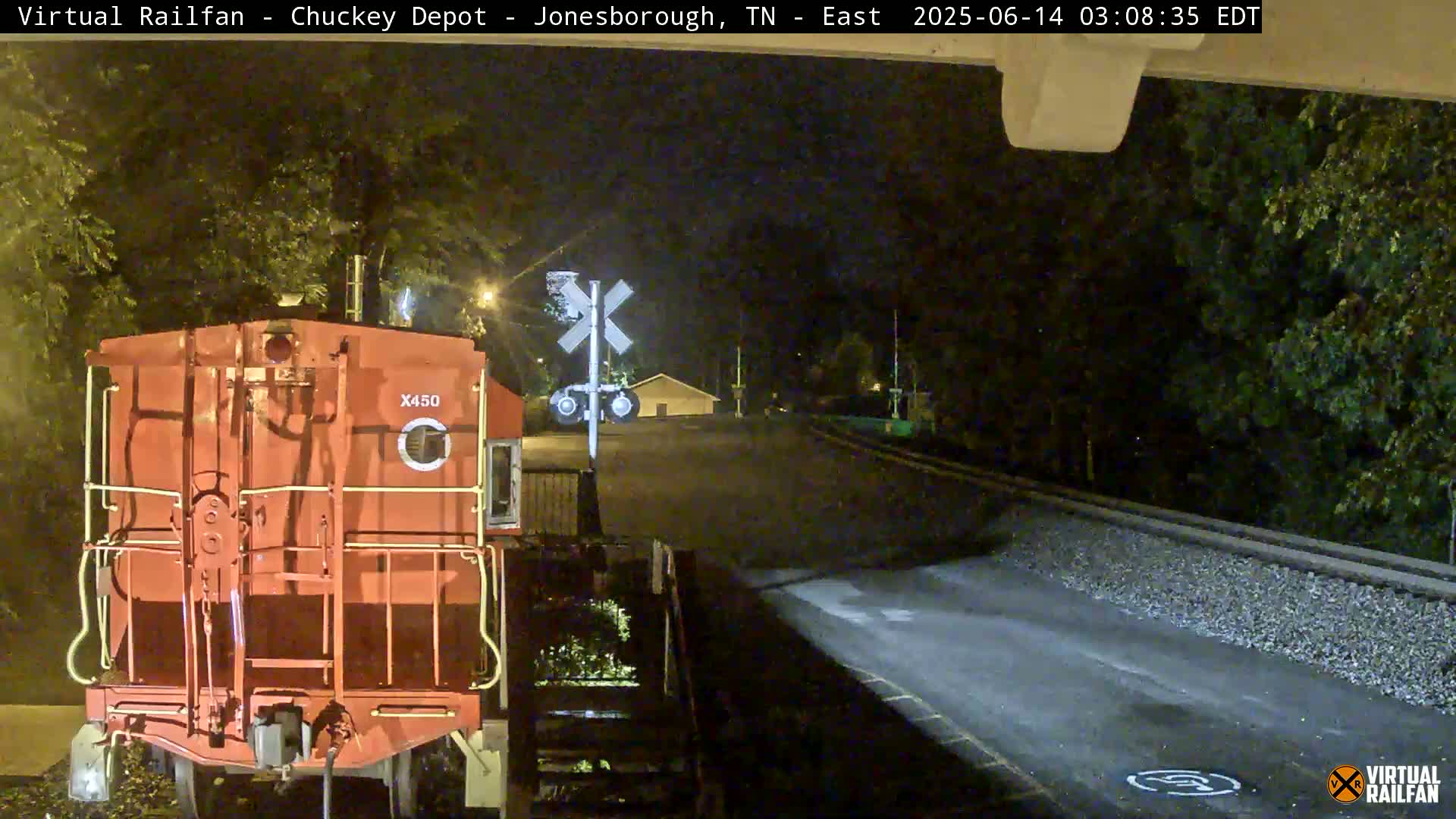 At night, a red train car sits at a railroad crossing near a roadway, under a clear sky.