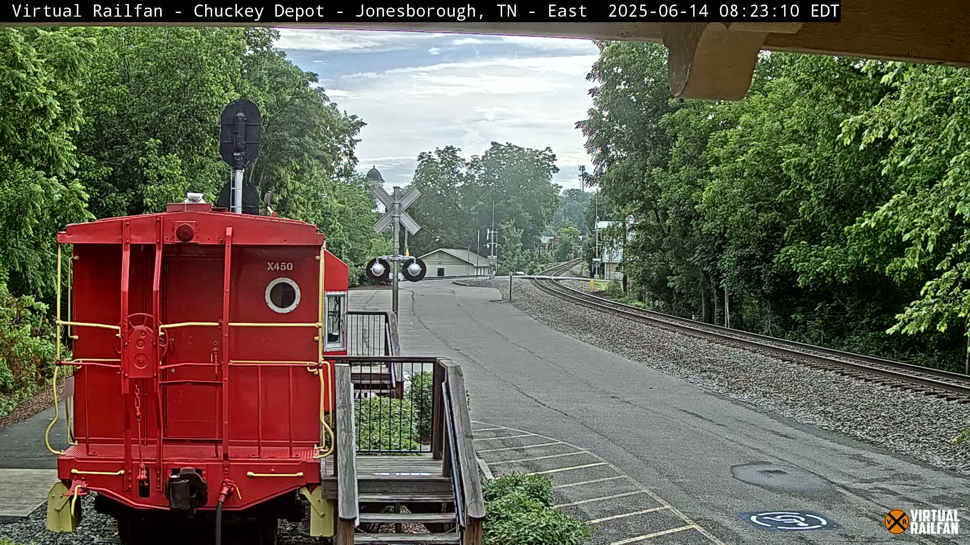 A red railroad car sits in front of a railway crossing, with a paved road leading to a curve in the tracks, all under a partly cloudy sky.