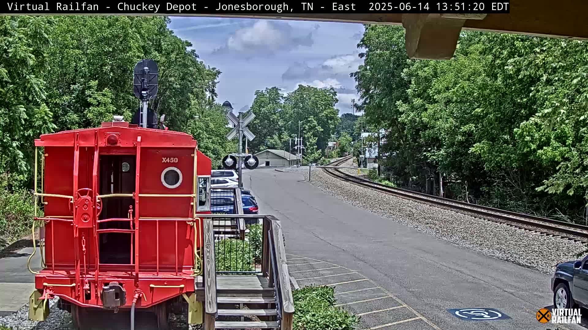 A red train car is parked facing the viewer, with a railway track and a paved road stretching away under a partly cloudy sky.