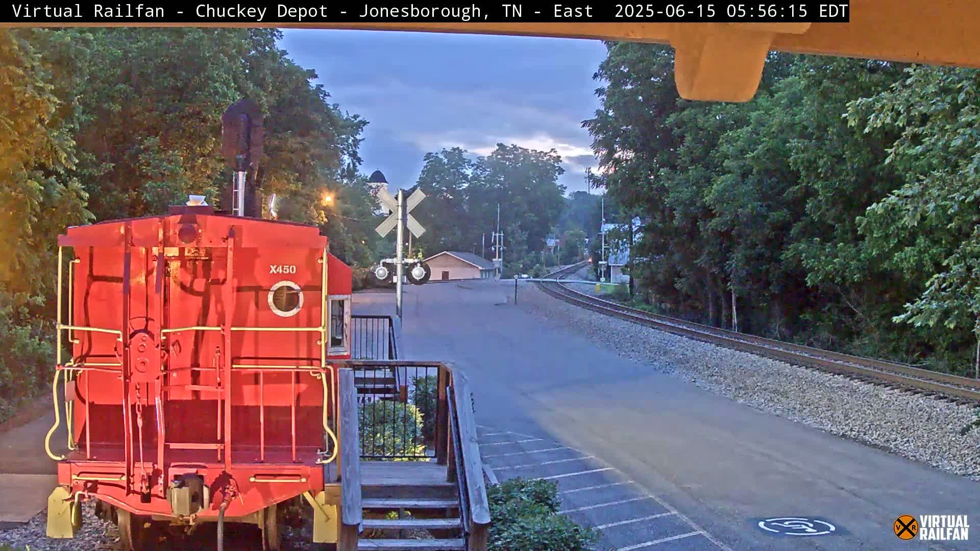 A red train caboose sits near a railroad crossing at dusk, with trees lining the track and a paved road leading toward a small building in the distance.