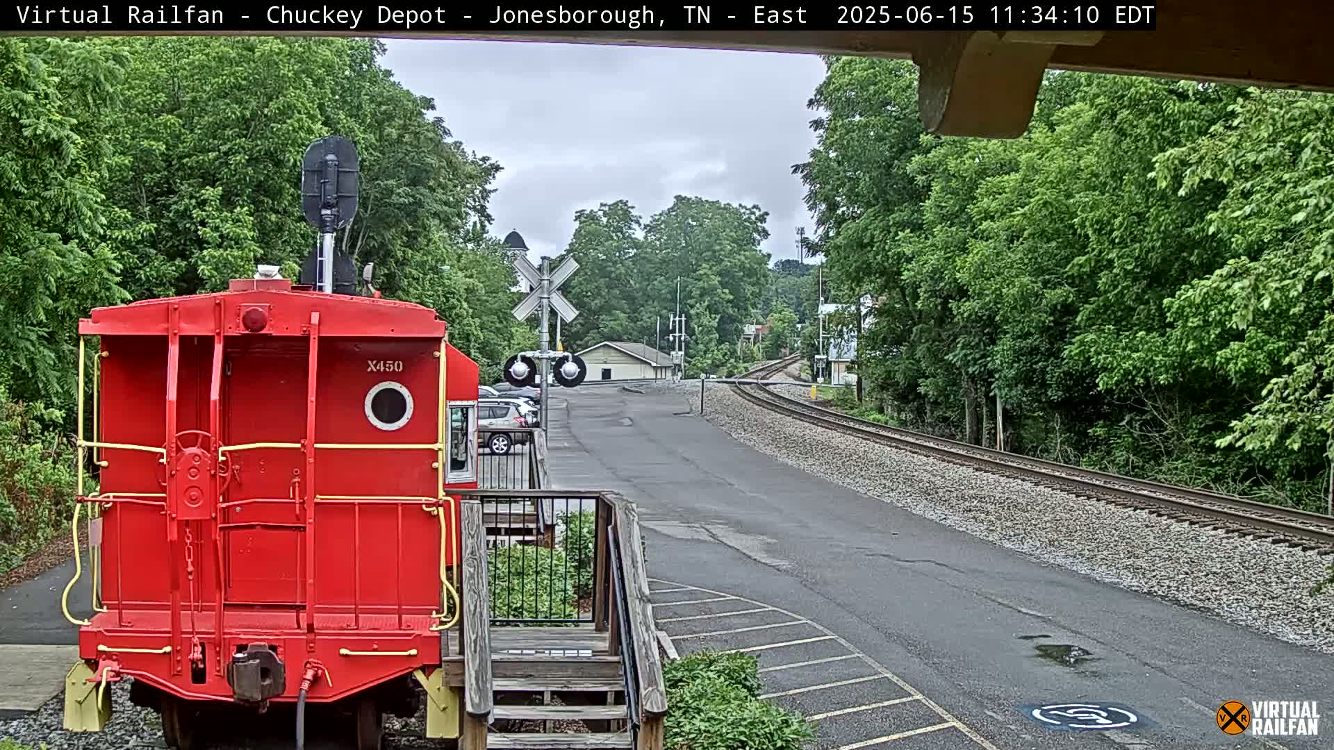 A red railway car is parked near a railway track that curves away into a tree-lined area under an overcast sky.