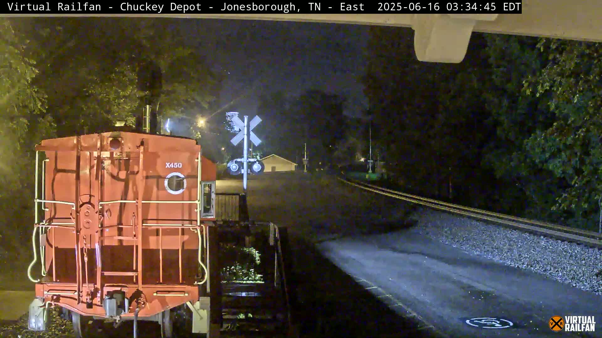 At night, a red train car is stopped at a railroad crossing near trees under a dark sky.