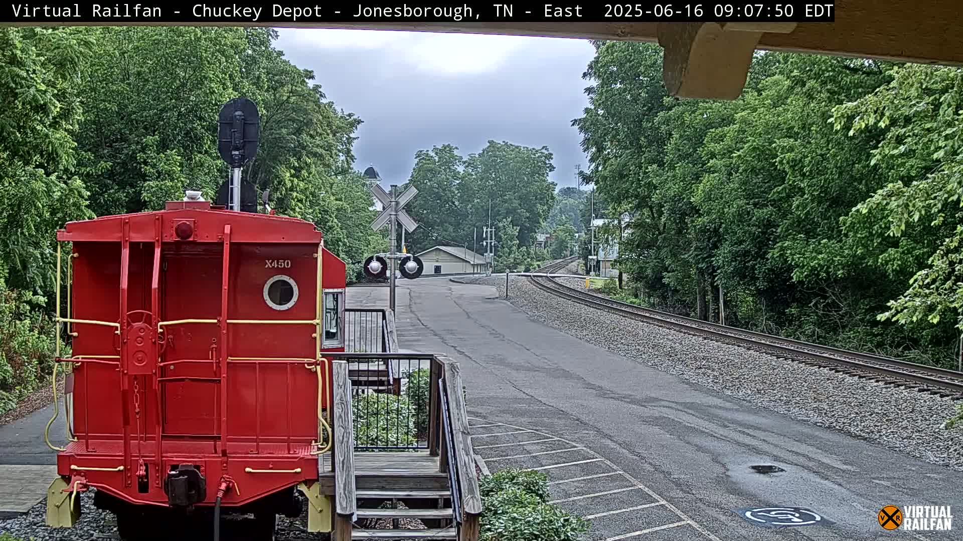 A red railroad car sits in front of a train track that curves into a tree-lined area on an overcast day.