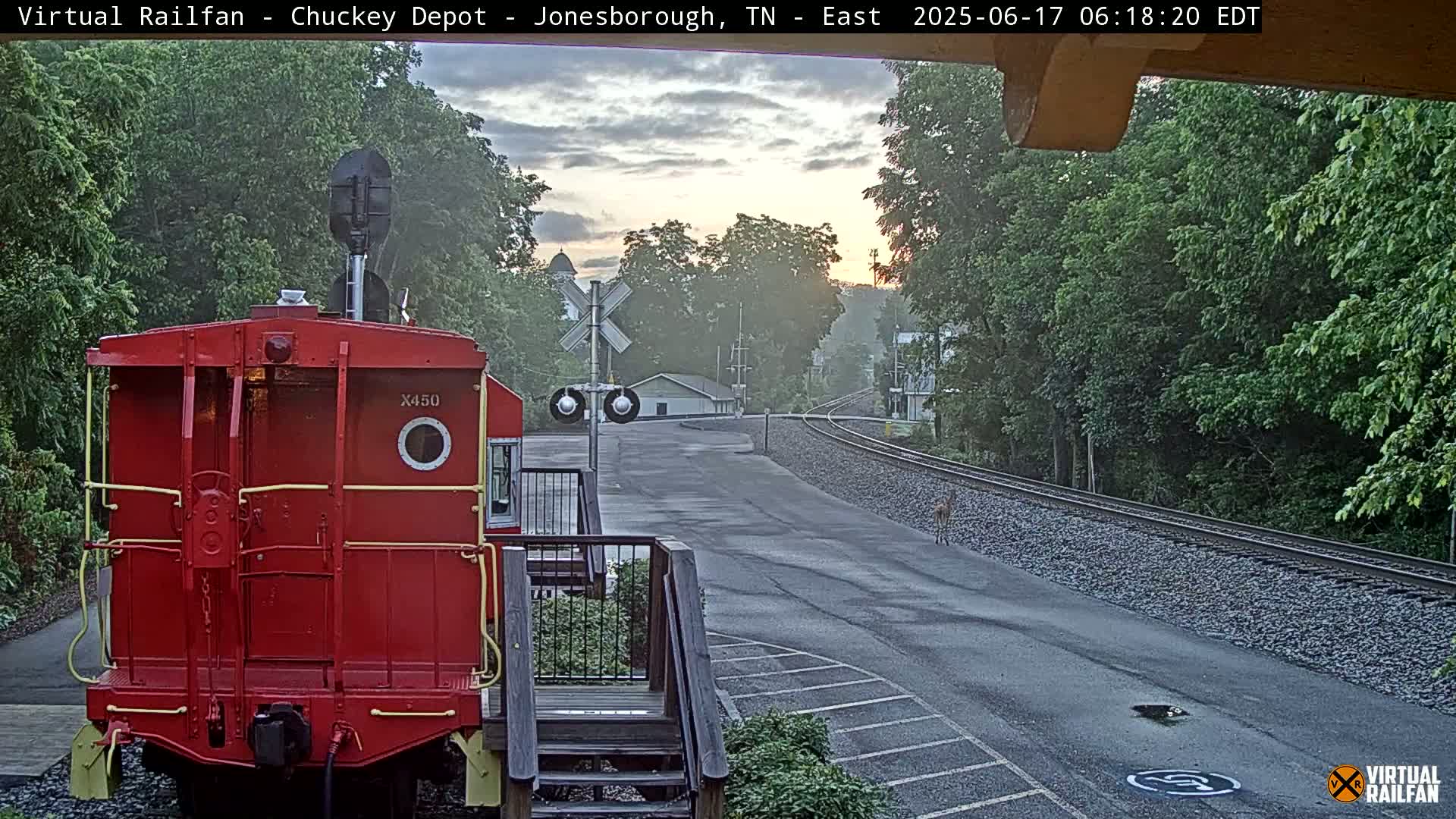A red railroad car sits in front of a railroad crossing, with a deer visible on the tracks in the distance, at sunrise under partly cloudy skies.