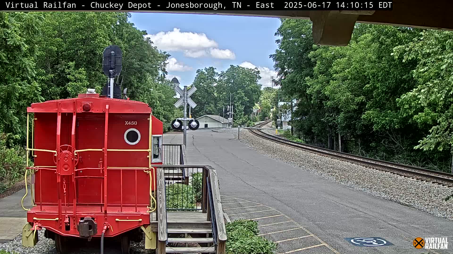 A red caboose sits near railroad tracks on a partly sunny day, with trees and a small building visible in the background.