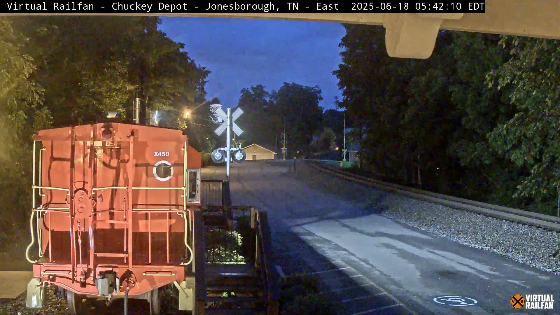 A red railroad car is parked at night near a railroad crossing under a twilight sky.