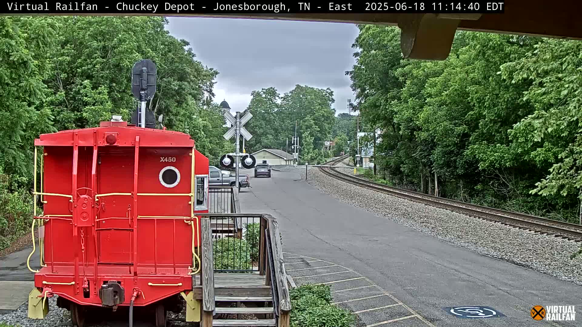 A red caboose is parked next to a railway track, with a road and a car visible in the distance under overcast skies.
