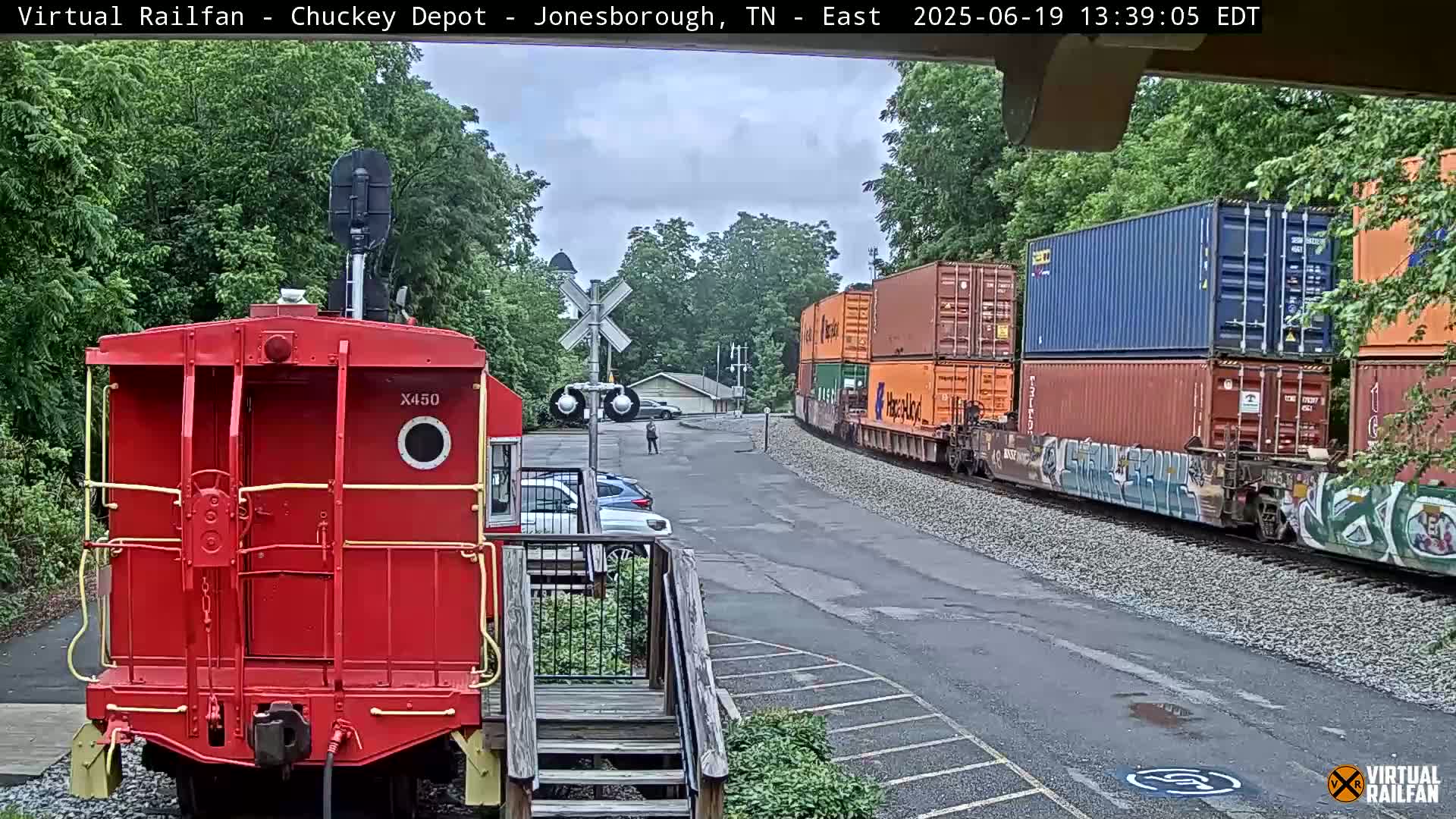 A red railcar is parked in front of a train carrying multiple shipping containers on a partly cloudy day.