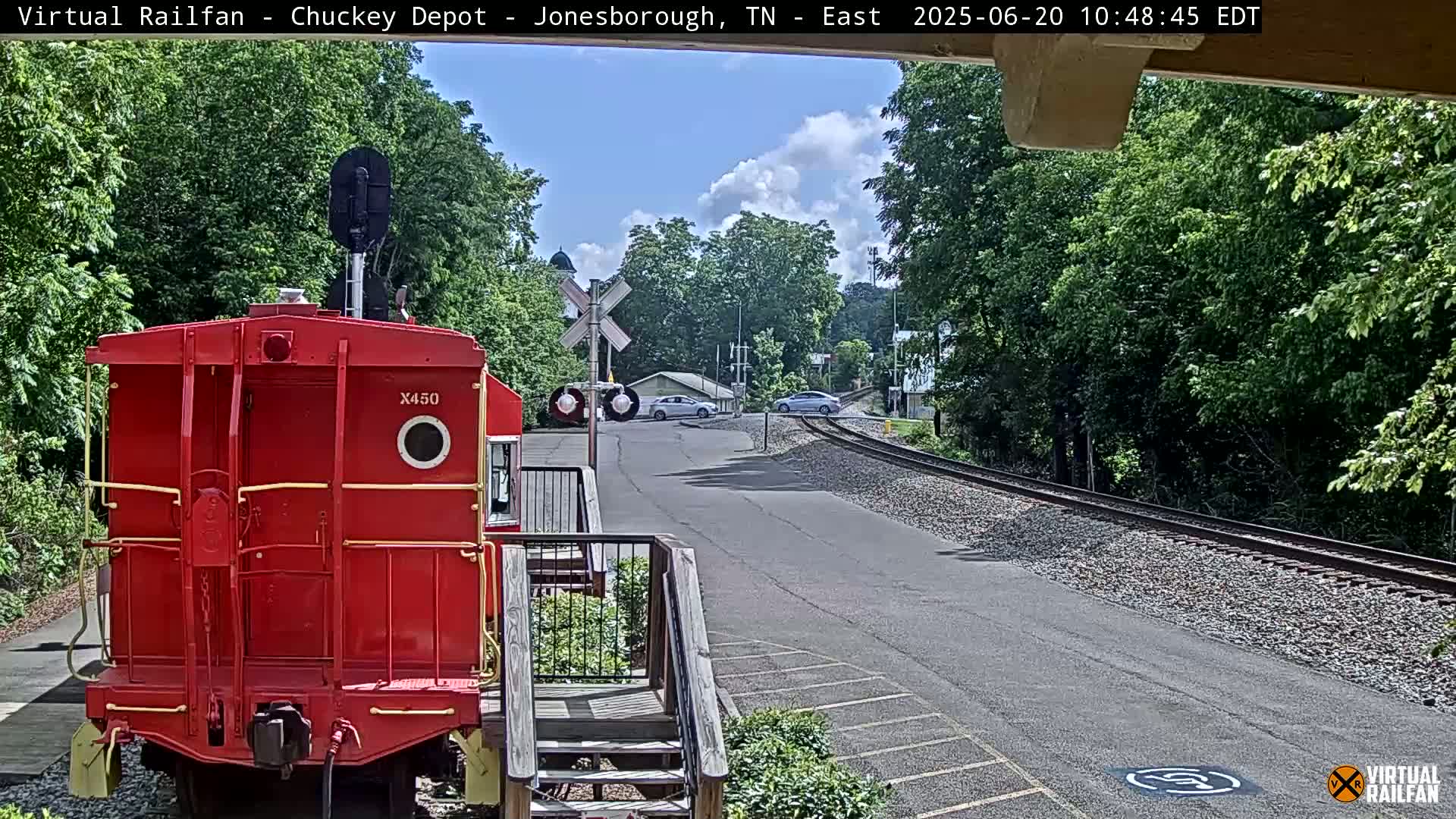 A red train car is parked near a railroad crossing on a partly cloudy day, with cars visible in the distance.
