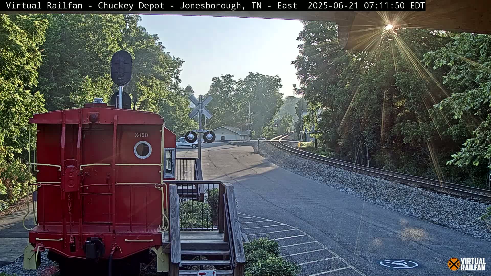 A red railroad car sits at a sunny train crossing next to a paved road, with trees and a building in the background.