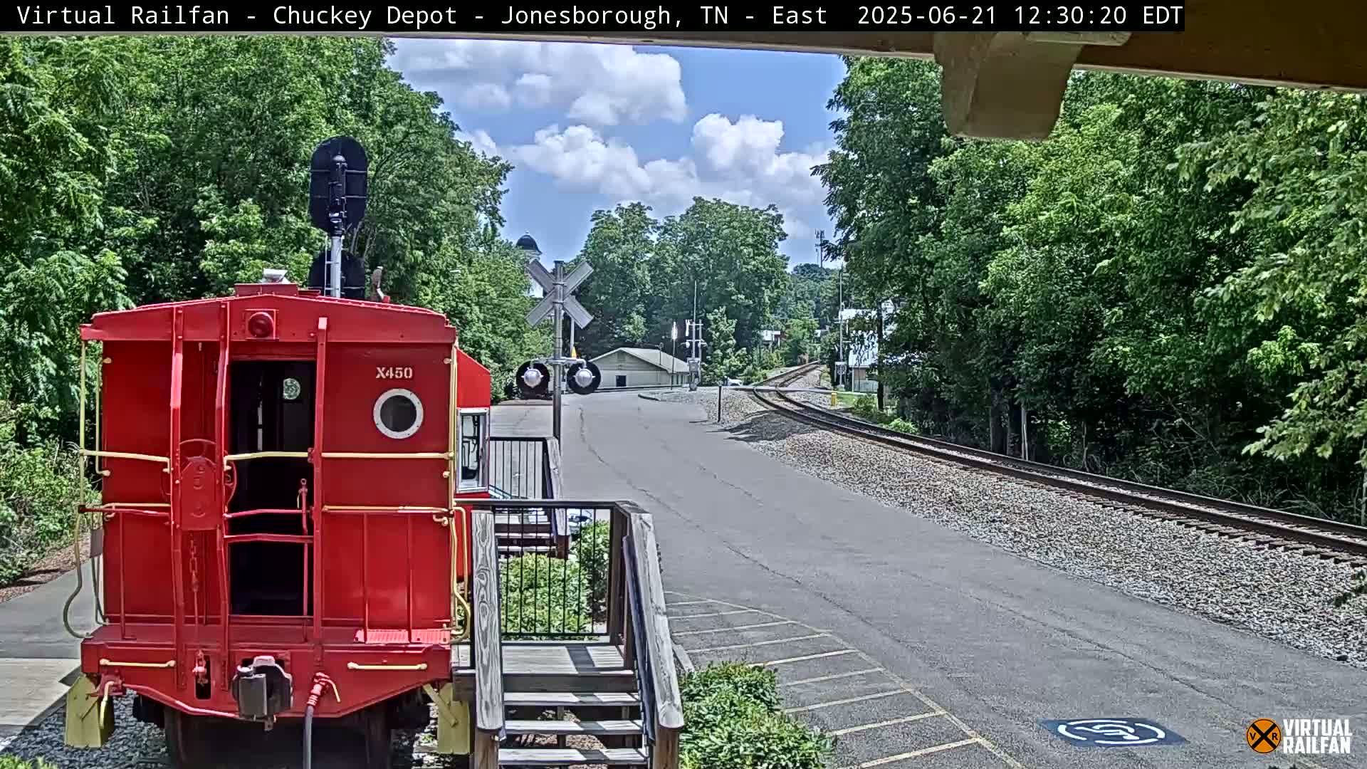 A red caboose sits in front of a railway crossing on a sunny day, with green trees and a paved road leading to a small building in the distance.