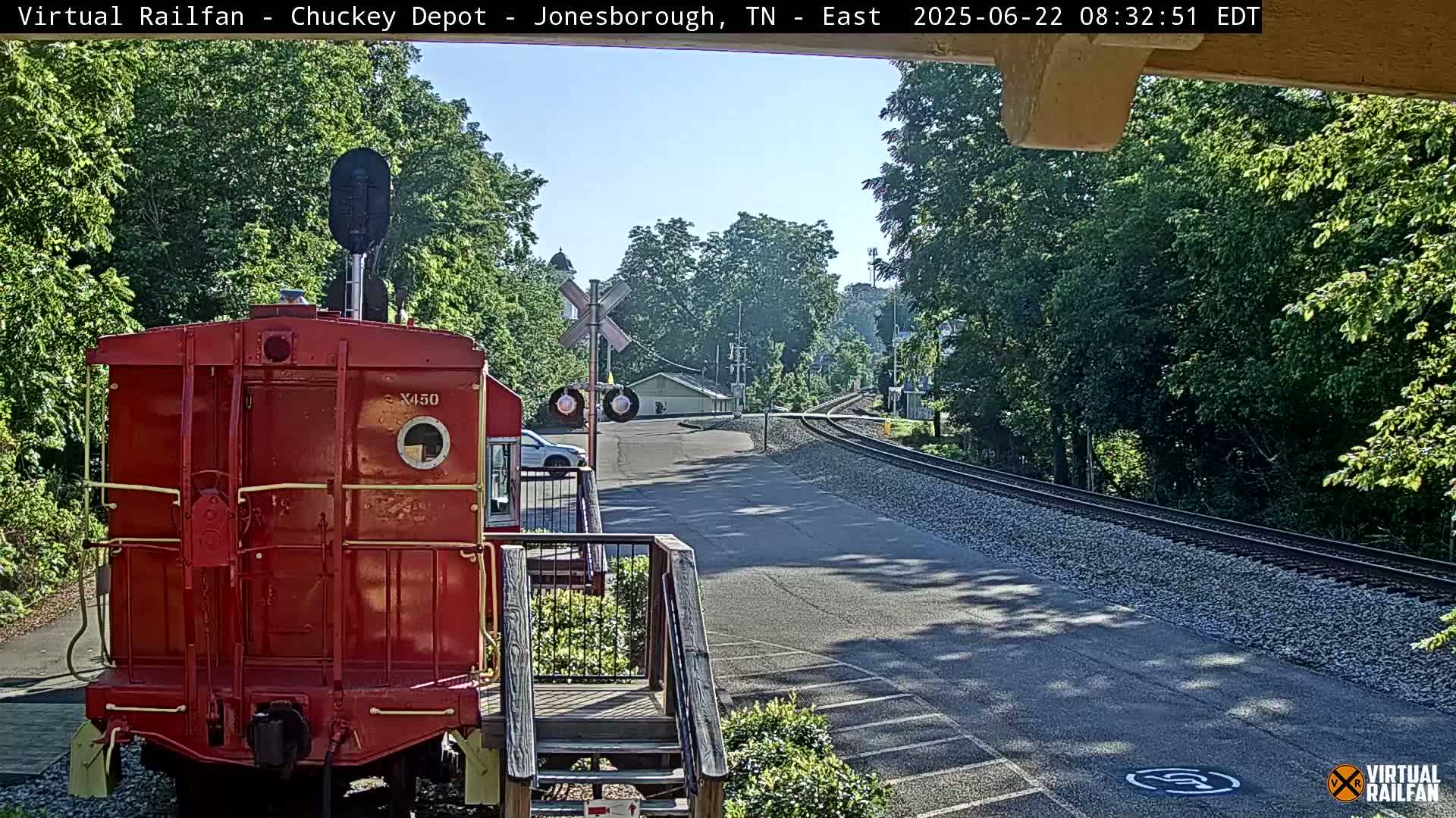 A red railroad car is parked at a railway crossing on a sunny day, with trees and a road visible in the background.