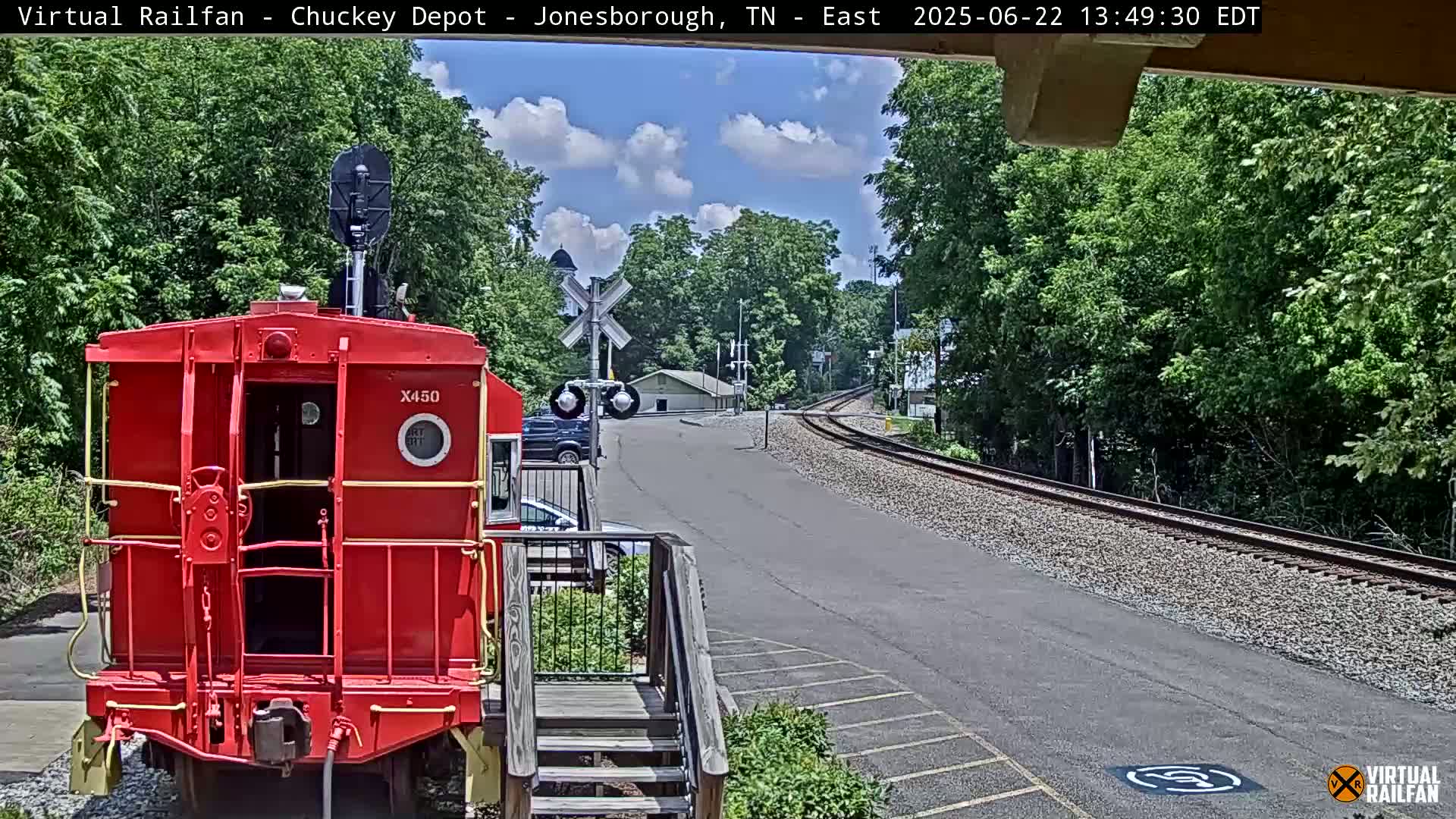 A red caboose sits near a railway track on a sunny day with lush green trees surrounding the area.