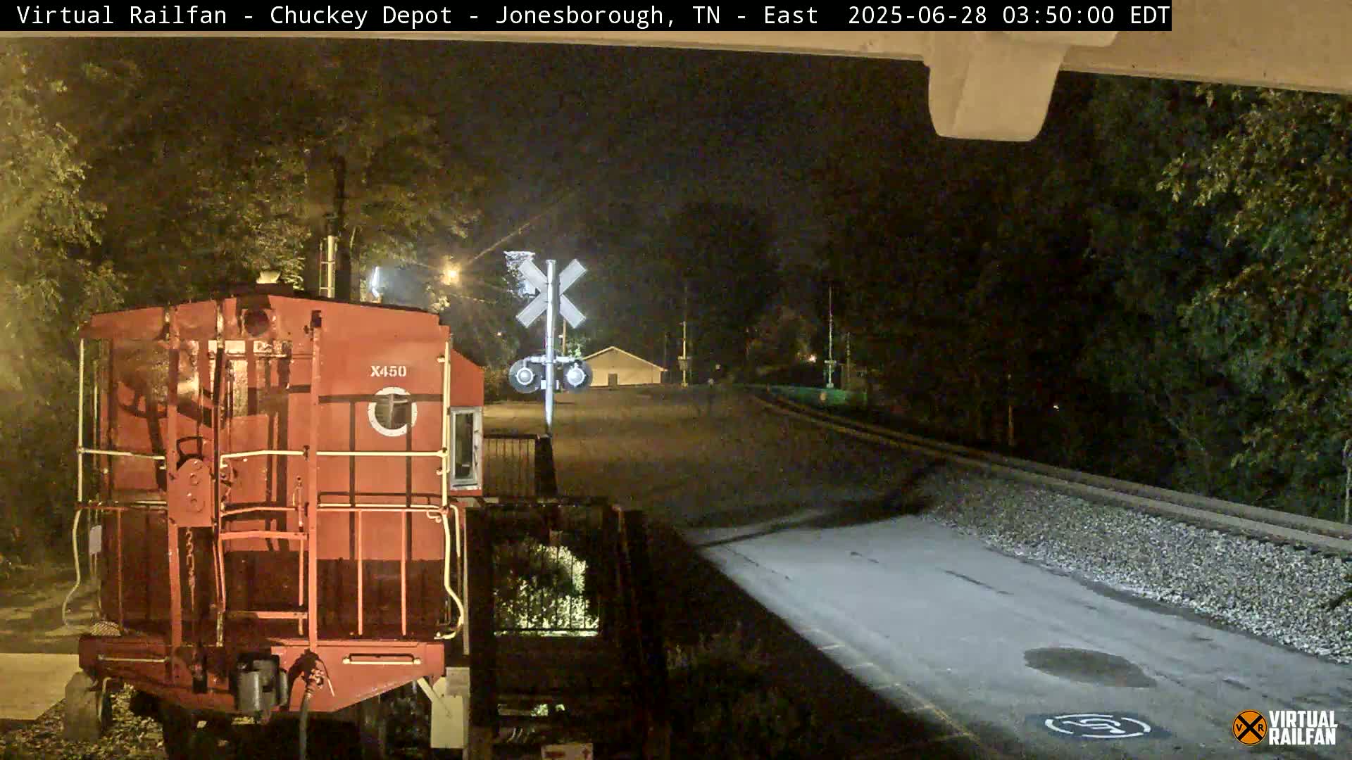 A red railroad caboose sits at a nighttime rail crossing under a dark sky.