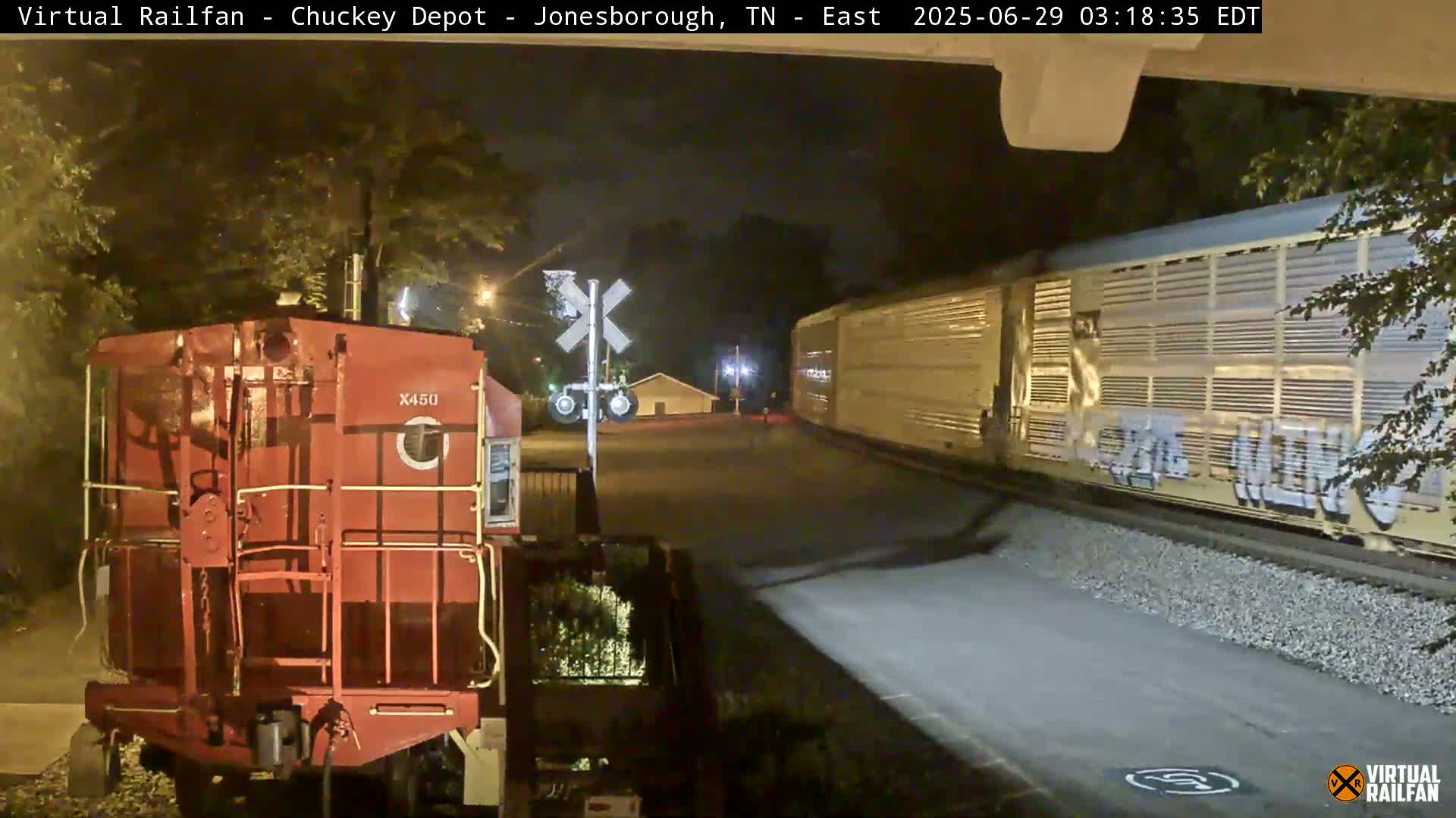 A red train car sits at a nighttime railroad crossing near a long freight train, illuminated by nearby streetlights.