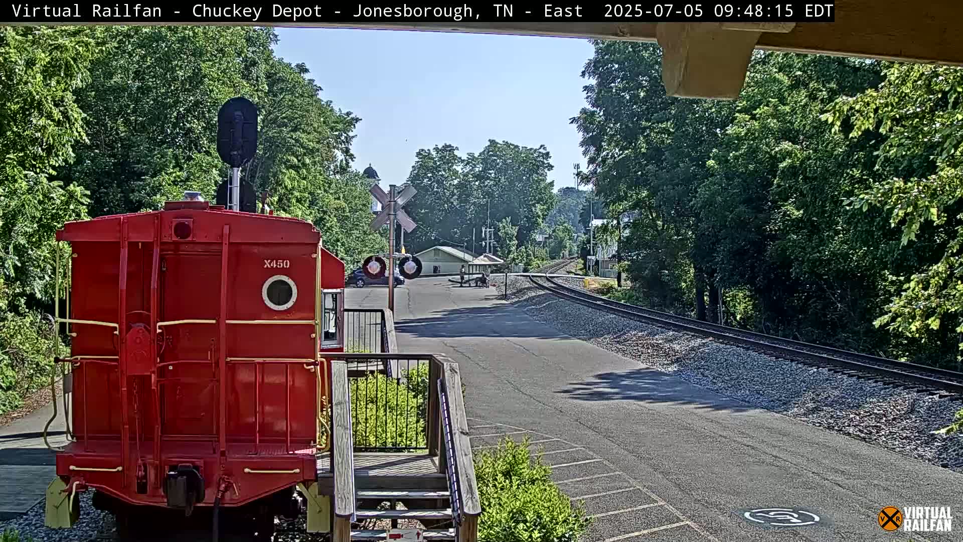 A red railway car is parked in front of a small train station on a sunny day, with train tracks extending into the distance amidst green trees.