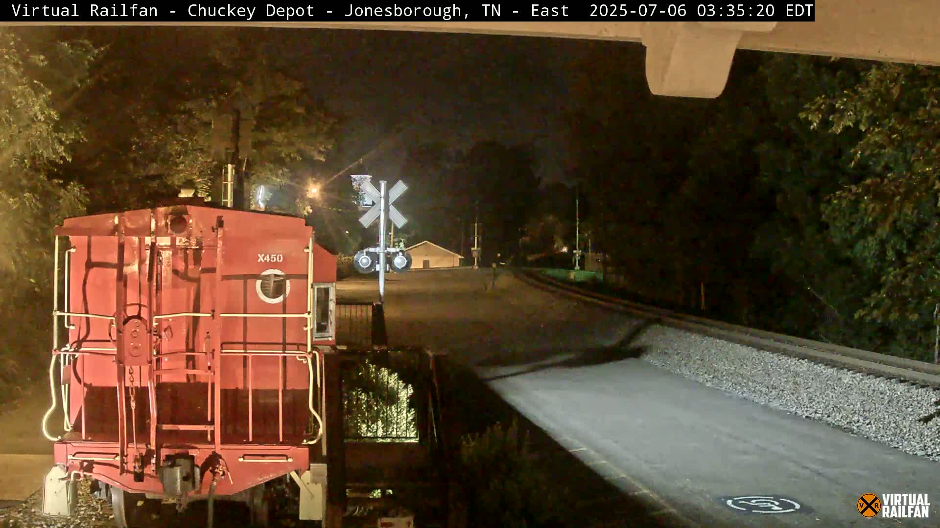 A red train car is visible at night, parked near a railroad crossing with a small building in the background.