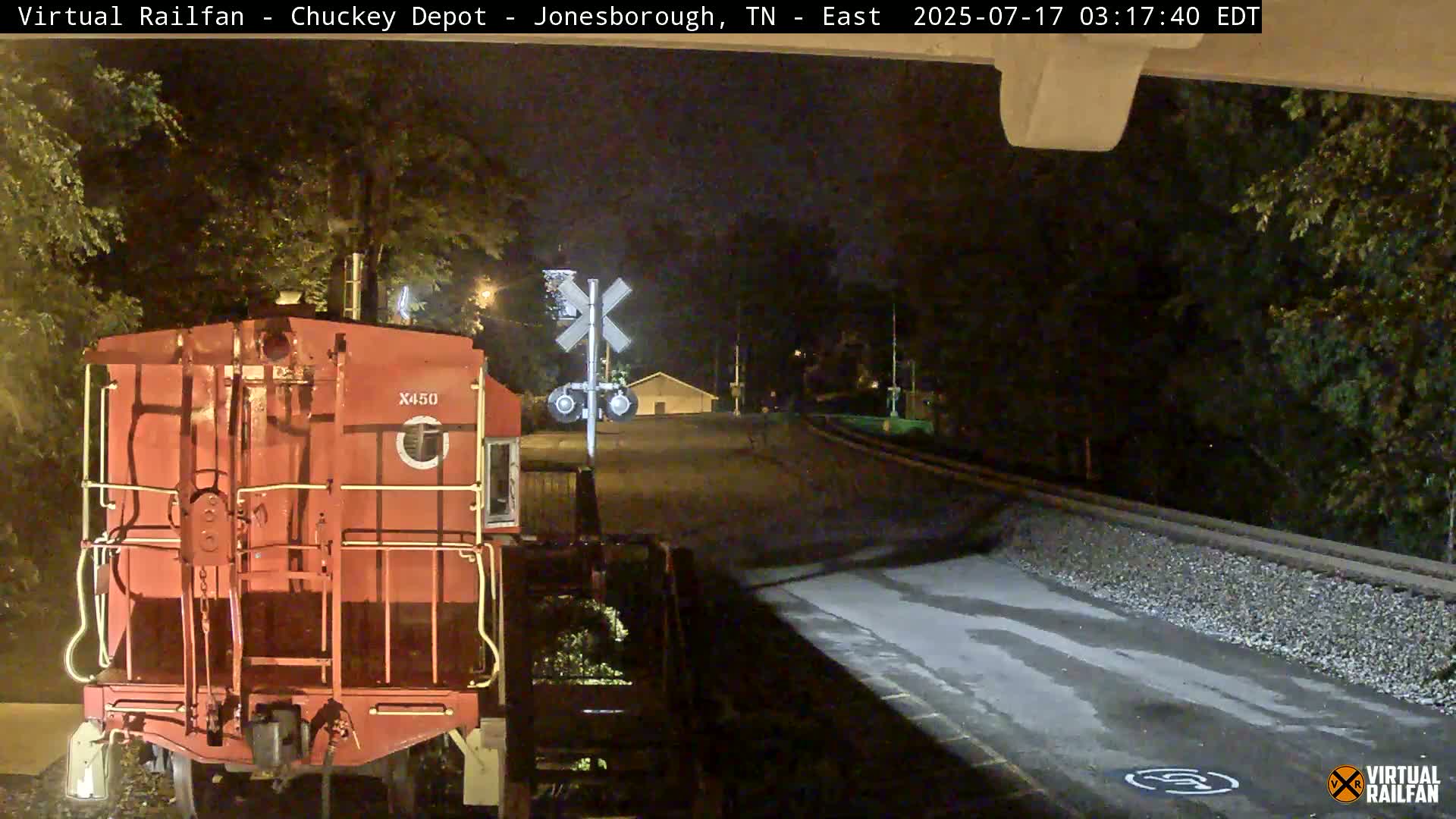 A red railroad car is parked at night near a railroad crossing, with trees and a road visible in the background.