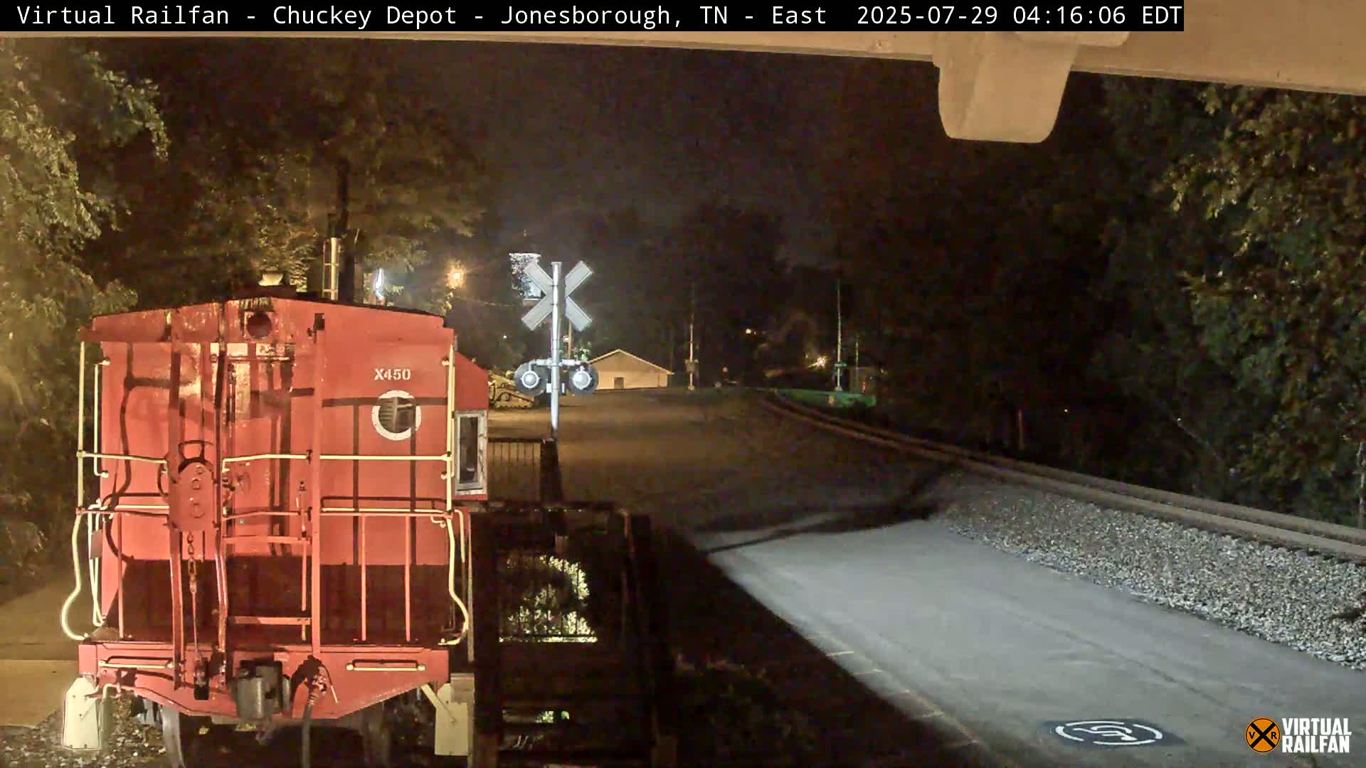 A red railroad car sits at a nighttime railroad crossing under a dark sky.