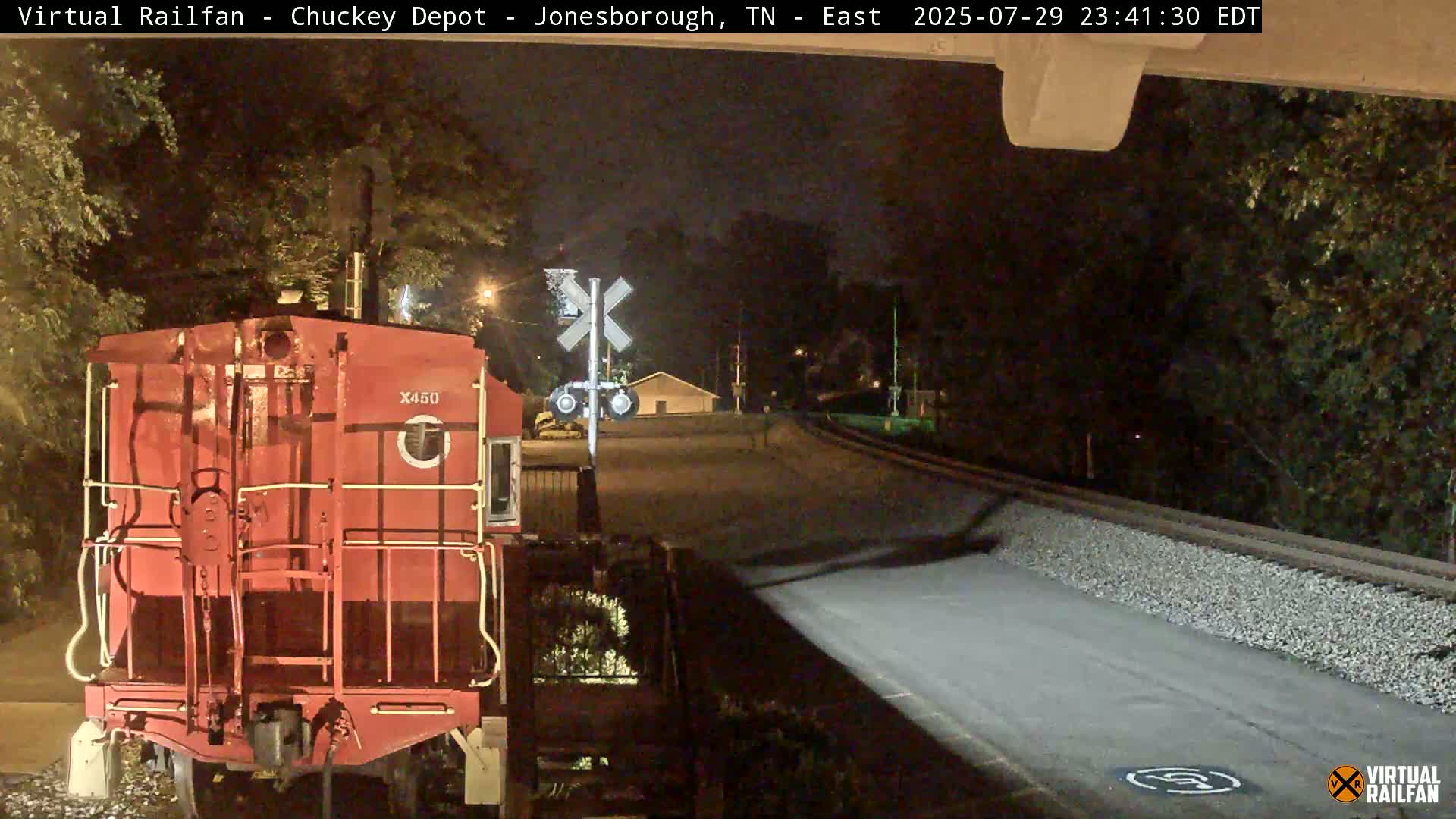 A red railroad car sits at a nighttime railroad crossing under a dark sky.